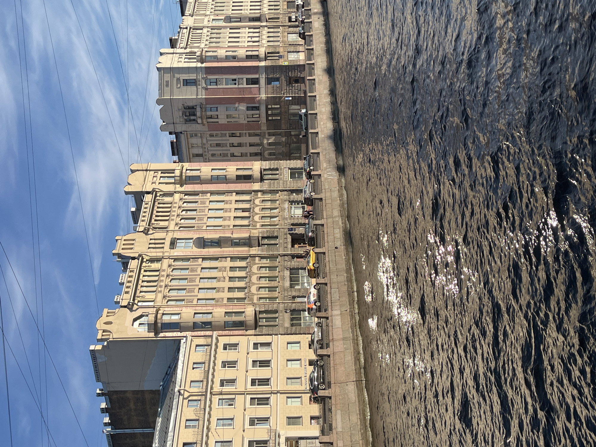 The image depicts a waterfront scene in Saint Petersburg, Russia. Multi-story buildings line the embankment, their facades exhibiting varying architectural styles and colors, ranging from pale yellow to faded brick. Cars are parked along the embankment directly in front of the buildings. The water has a dark, textured surface with sunlight reflecting off of it. Overhead, a web of power lines is visible against a blue sky with wispy clouds. No people are clearly discernible in the image.