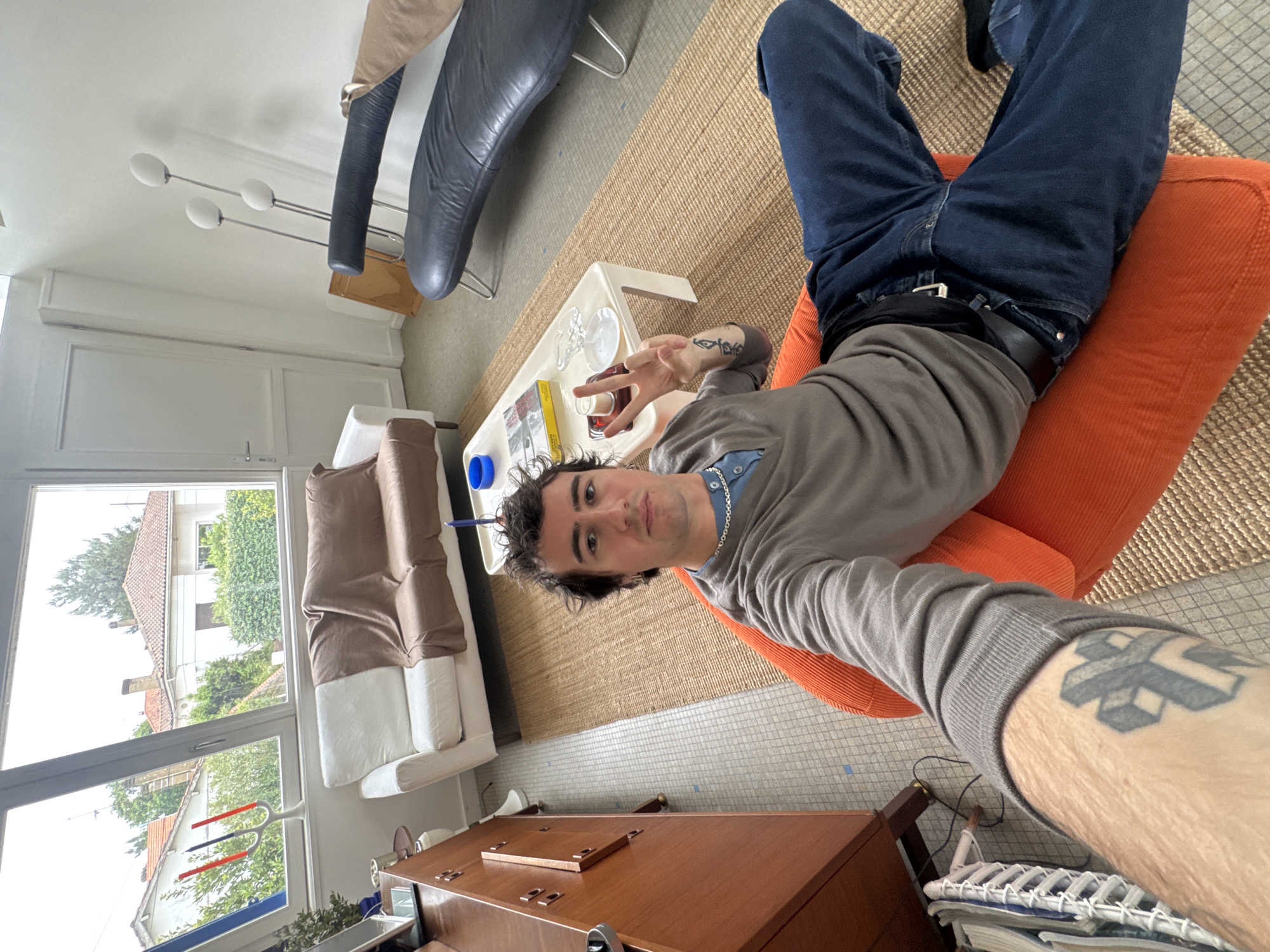 A man is lying on an orange chair in a room in Soulac-sur-Mer, France. The man is wearing blue jeans, a gray long-sleeved shirt, and a black belt. He is facing the camera and making a peace sign with his left hand. He has short, dark hair and a silver chain around his neck. The room contains a white couch with brown cushions, a wooden cabinet, a black leather chair, a white coffee table, and a tan rug. Outside the window, trees and buildings are visible. The floor is light gray tile. The photo appears to be taken with a wide-angle lens, resulting in some perspective distortion. The lighting is bright, suggesting it is daytime. The man has tattoos on his arms.