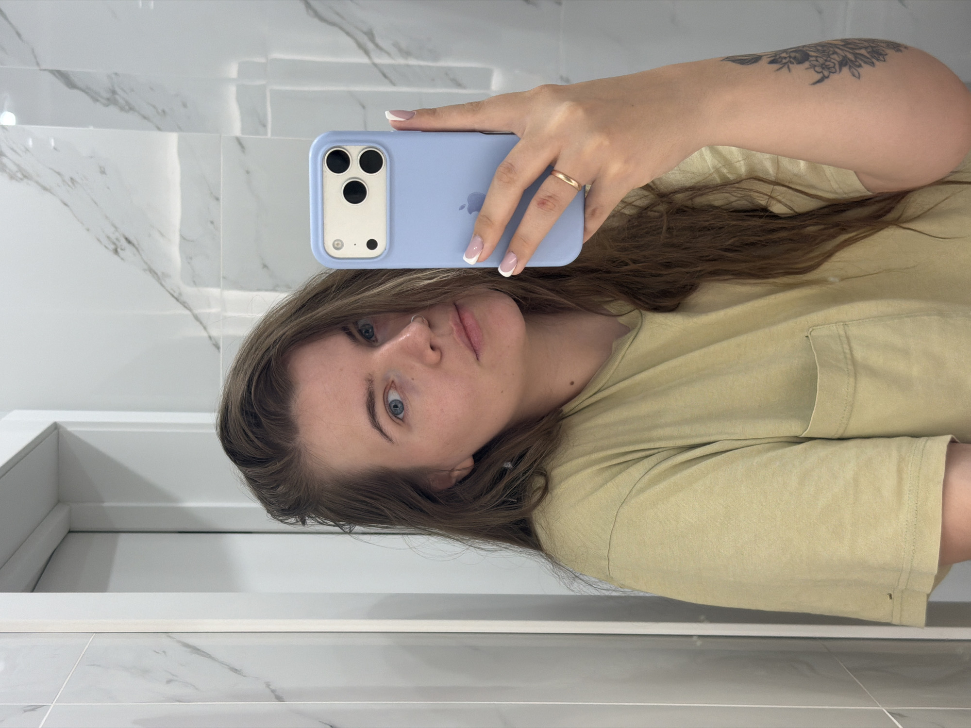 A young woman is taking a selfie in a bathroom with white marble-like tile. She is wearing a light yellow t-shirt and has shoulder-length brown hair. She is holding a blue iPhone with a triple camera setup. She has a gold ring on her finger and a tattoo on her upper arm. The location is unspecified, therefore the city of Chaikovsky, Russia, cannot be verified.
