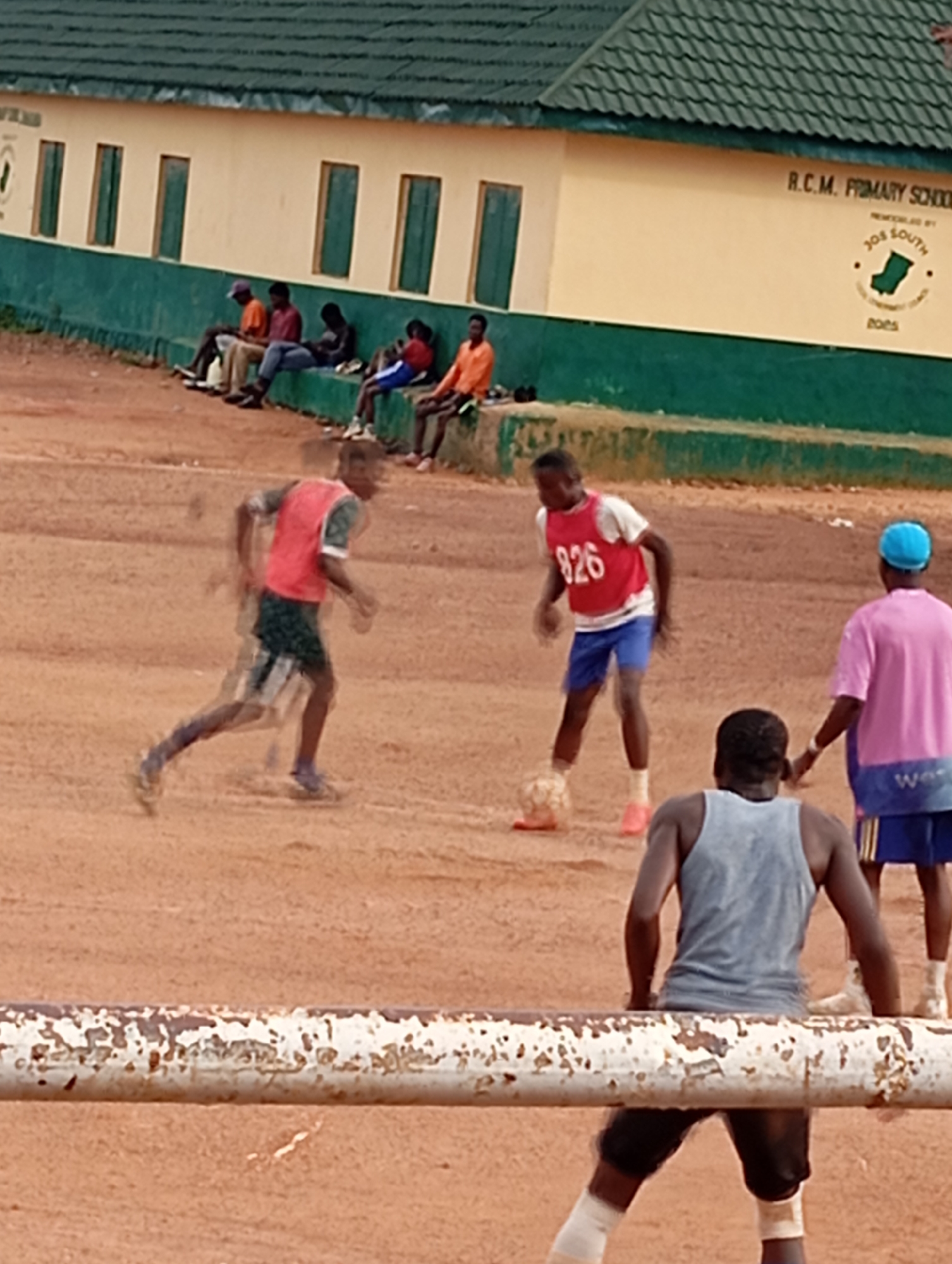 This media file depicts an outdoor scene at R.C.M. Primary School in Jos, Nigeria. The primary subjects are several young people engaged in a game of football on a dusty, reddish-brown field. Two individuals are actively playing, one wearing a red vest with the number "826" and blue shorts, and the other in a red and white shirt and green shorts, captured in motion blur. A third person in a pink t-shirt and blue shorts with yellow stripes is observing, and another person in a grey tank top and black shorts is positioned in the foreground, seemingly waiting to join the game.
In the background, a light yellow school building with green trim and several windows runs along the top of the frame. Text visible on the building reads "R.C.M. PRIMARY SCHOOL" and a circular logo with "JOS SOUTH" and what appears to be a map of Nigeria. The overall impression is of a casual, perhaps after-school, football match. The lighting suggests it is daytime, and the dusty ground indicates dry weather conditions. The people are dressed in casual sportswear, and their postures and the dynamic motion blur suggest an active and energetic event. The presence of spectators and players suggests a community or school sporting activity.