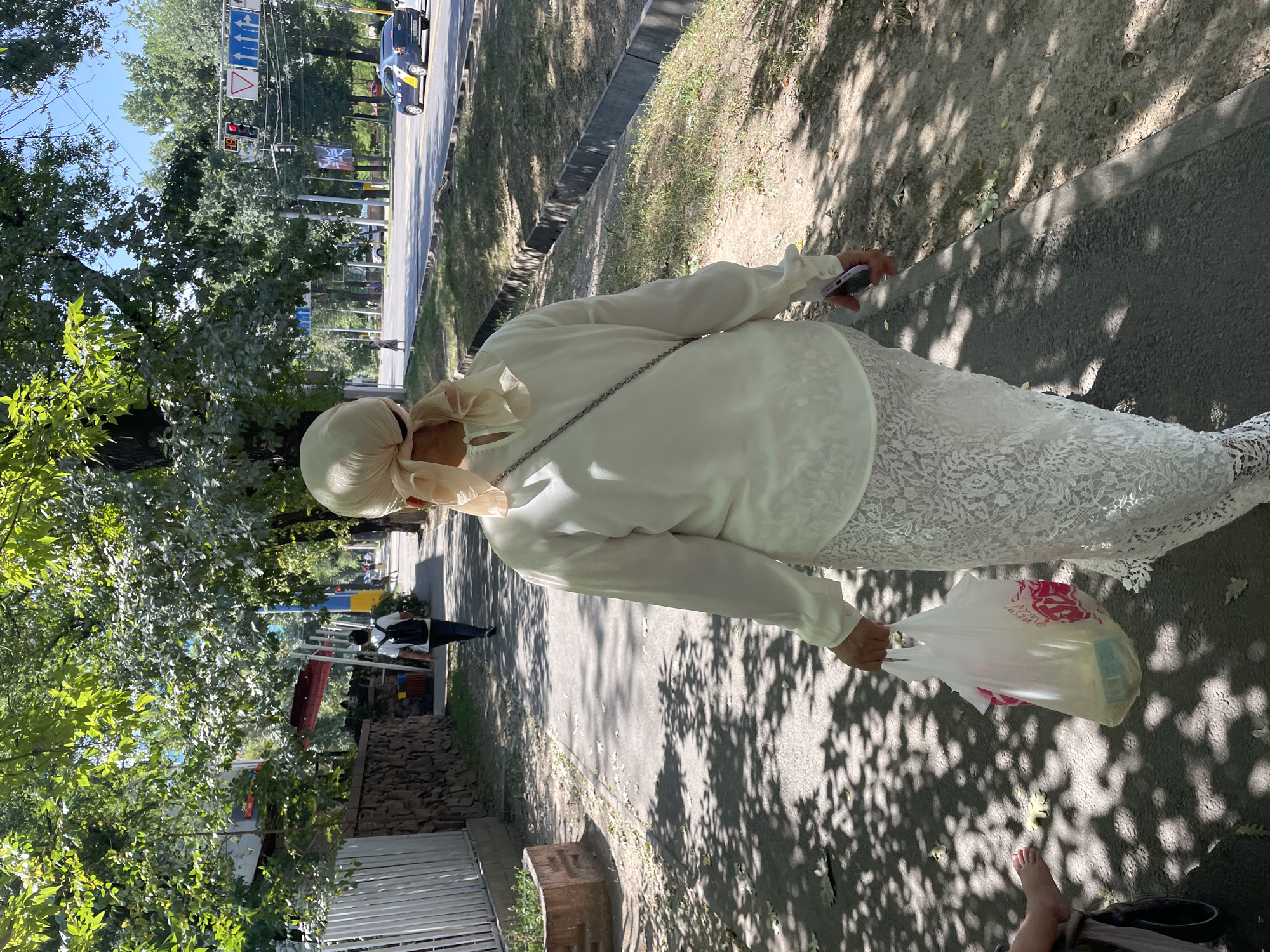 A woman in a white blouse and white lace skirt walks away from the camera on a tree-lined sidewalk in Almaty, Kazakhstan. She wears a light beige headscarf tied at the back. She holds a white plastic shopping bag with pink lettering in her right hand and a purple cellphone in her left. Her left foot is visible, bare, in what appears to be a stroller to the left of the frame. The sidewalk is paved and dappled with sunlight and shadows from the trees. A stone wall with white fencing runs along the left side of the sidewalk. Across the street, there is a road with traffic, including a yellow and blue taxi, and pedestrian signals. The environment suggests a sunny day in a developed urban area with significant greenery.
