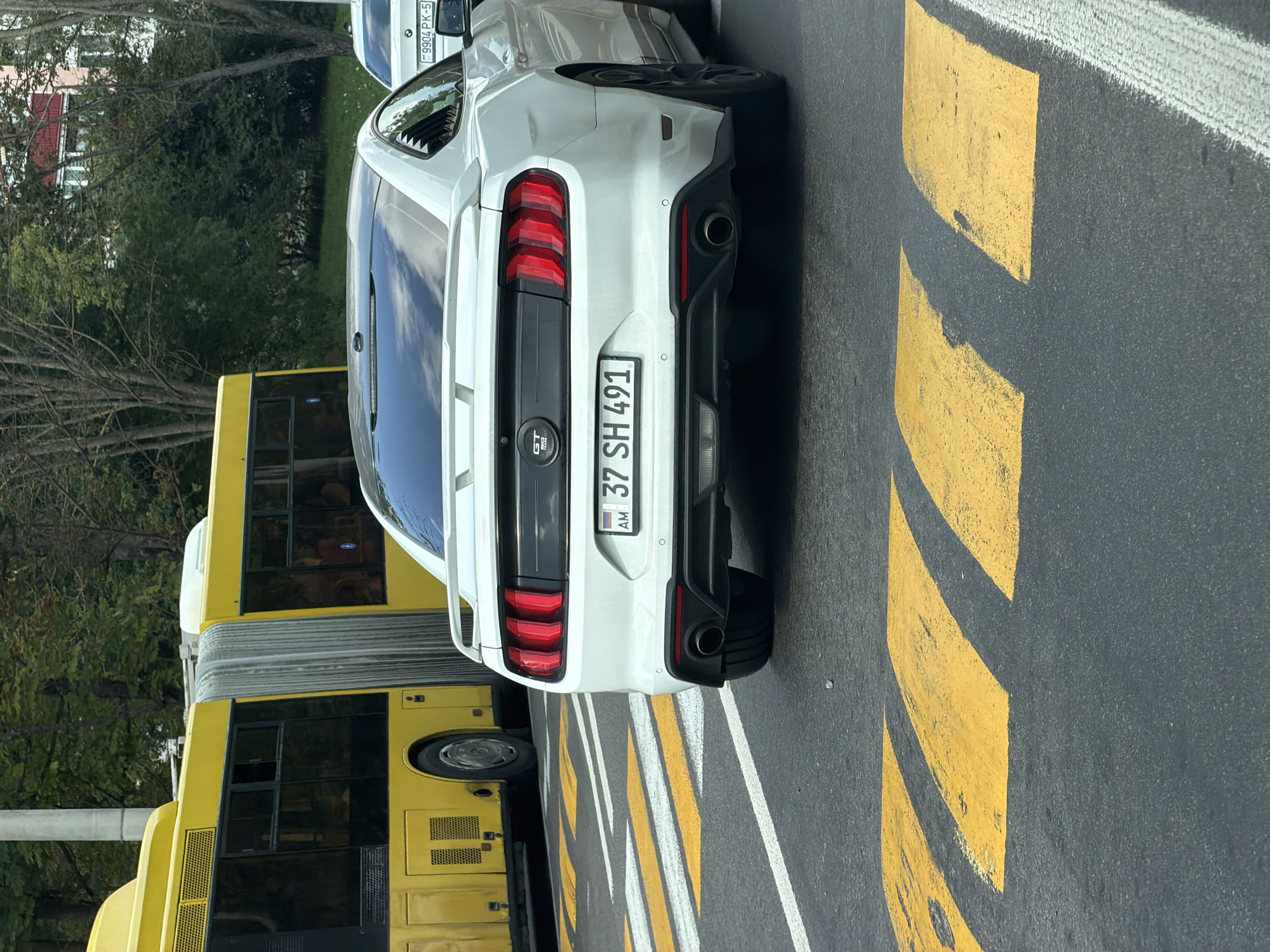 The image shows the rear of a white Ford Mustang in Minsk, Belarus. The car has a license plate that reads "37 SH 491" and has an Armenian flag symbol. The Mustang is stopped at a crosswalk marked with yellow and white stripes. To the left of the Mustang, a yellow articulated bus is visible. The environment appears to be an urban setting with trees and buildings in the background. The image is taken from a low angle, looking towards the back of the car.