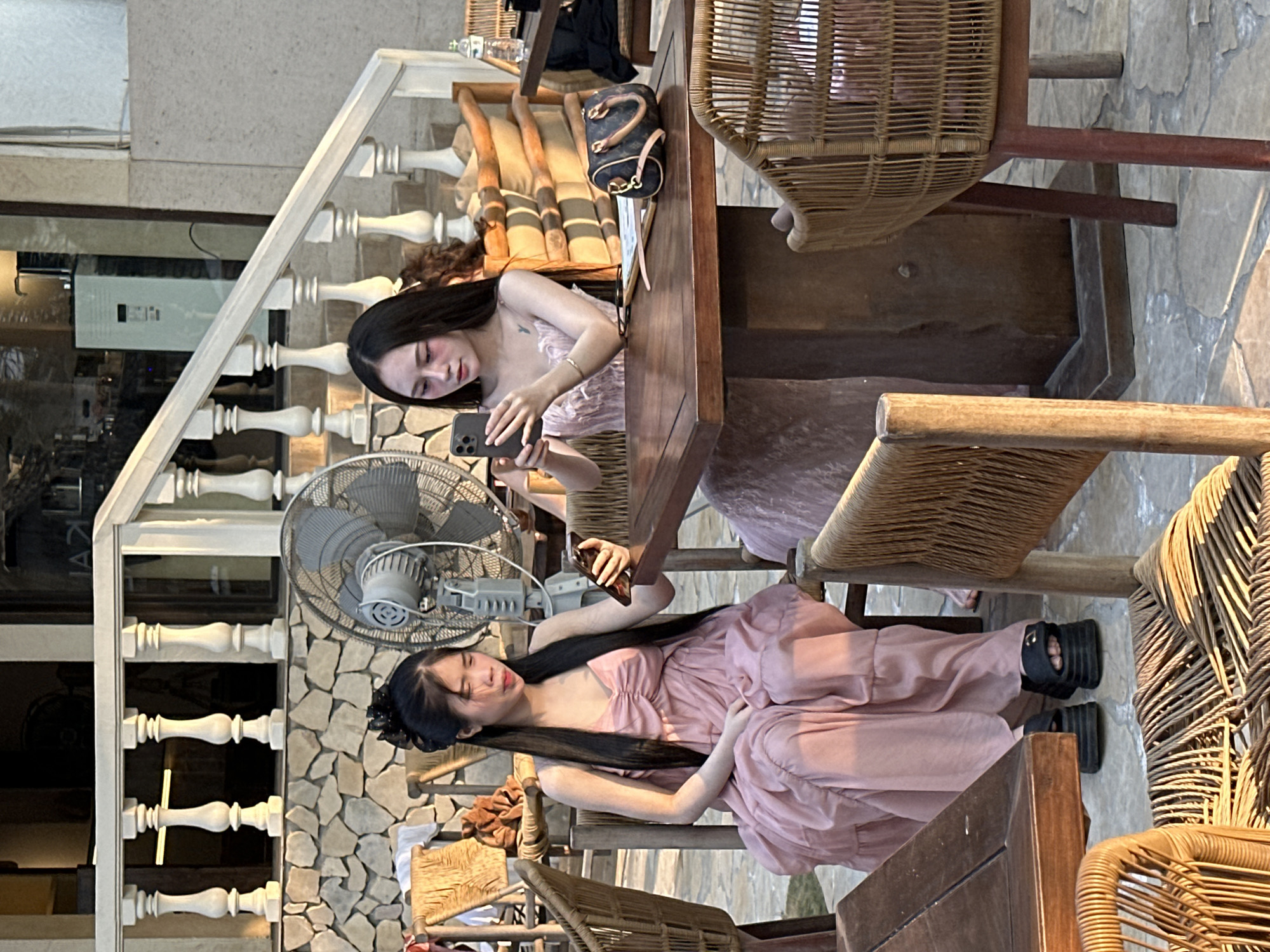 Two young women are seated at an outdoor table with wicker chairs in Đà Nẵng, Vietnam. One woman, wearing a light pink dress, reclines in her chair, while the other, also in a pink dress, is holding a smartphone and appearing to take a photo. A vintage-style electric fan is visible behind them, and a decorative staircase is partially seen in the background. A handbag rests on the table. The setting appears to be a restaurant or cafe.