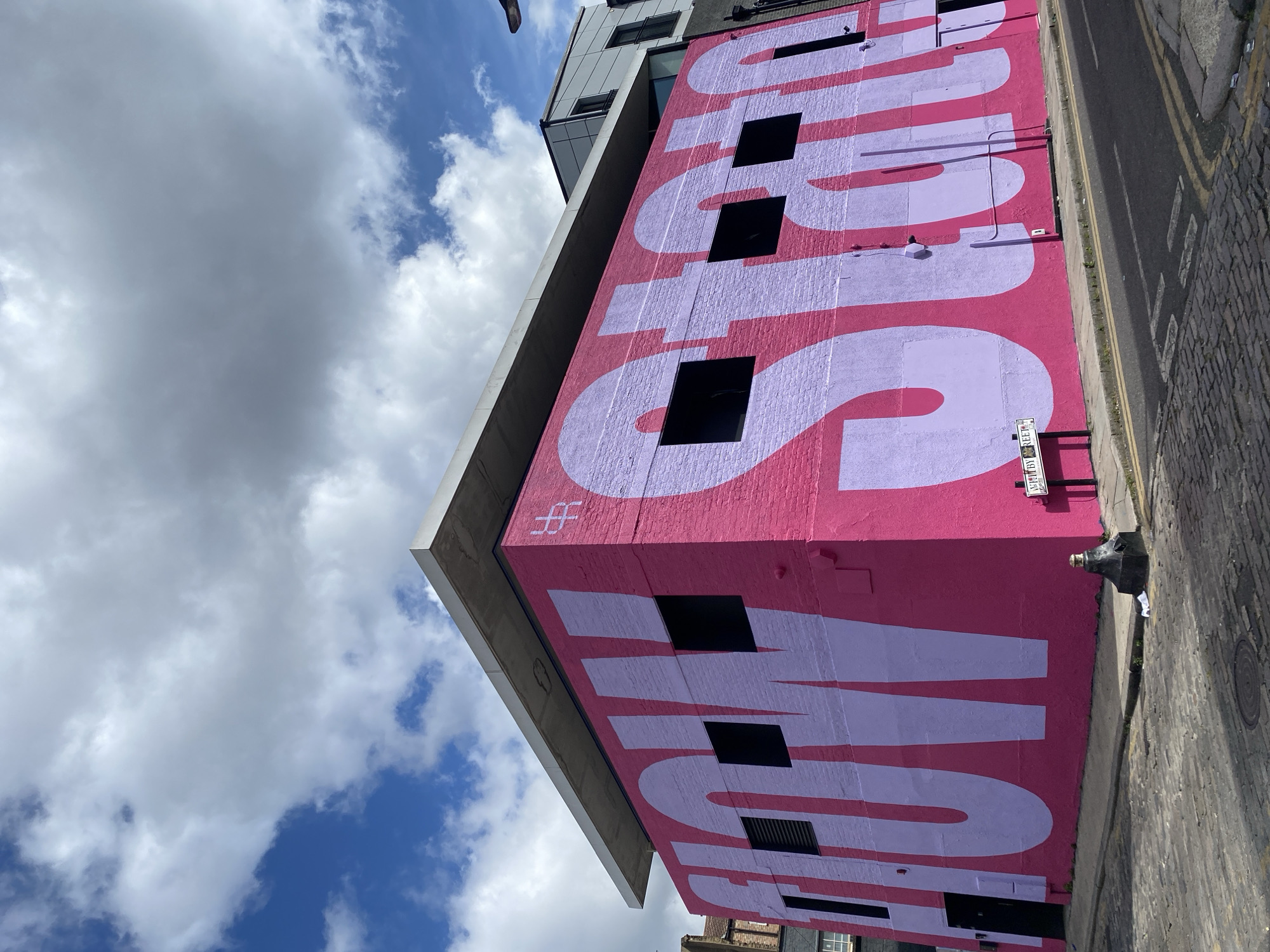 The image depicts the exterior of a building in London, United Kingdom, featuring a large pink mural with the word "SHOWstudio" painted in light purple. The building's facade incorporates windows into the design of the mural. A street sign for "Brewer Street" is visible on the side of the building. The sky is partly cloudy with patches of blue. A portion of the street and sidewalk can be seen at the bottom of the frame.