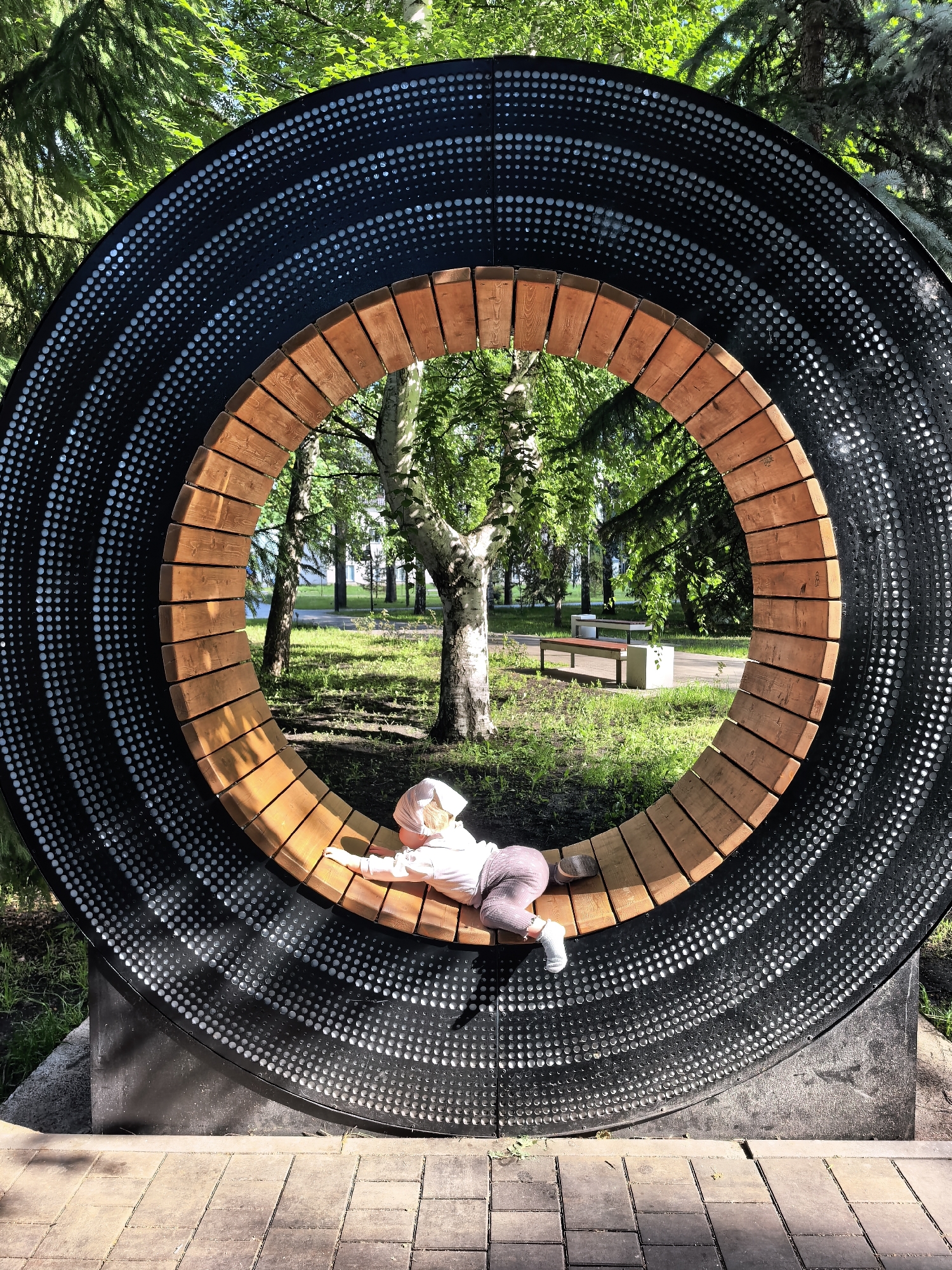 In Sterlitamak, Russia, a young child lies on their stomach on a curved wooden bench inside a large, circular structure resembling a stylized vinyl record. The outer rim of the "record" is black and perforated with a pattern of small, circular indentations, arranged in concentric circles and radial lines. The inner portion of the structure is formed by a semicircular bench made of light-colored wooden planks. The child, wearing a light-colored headscarf, a light pink shirt, and grey pants, is positioned near the center of the bench, facing towards the right side of the frame. Their left arm is extended forward, and their right arm is bent at the elbow. The background visible through the circular opening shows a park setting with green grass, mature trees with lush foliage, and paved pathways. In the distance, park benches can be seen. The sunlight casts shadows on the ground, indicating a bright day.