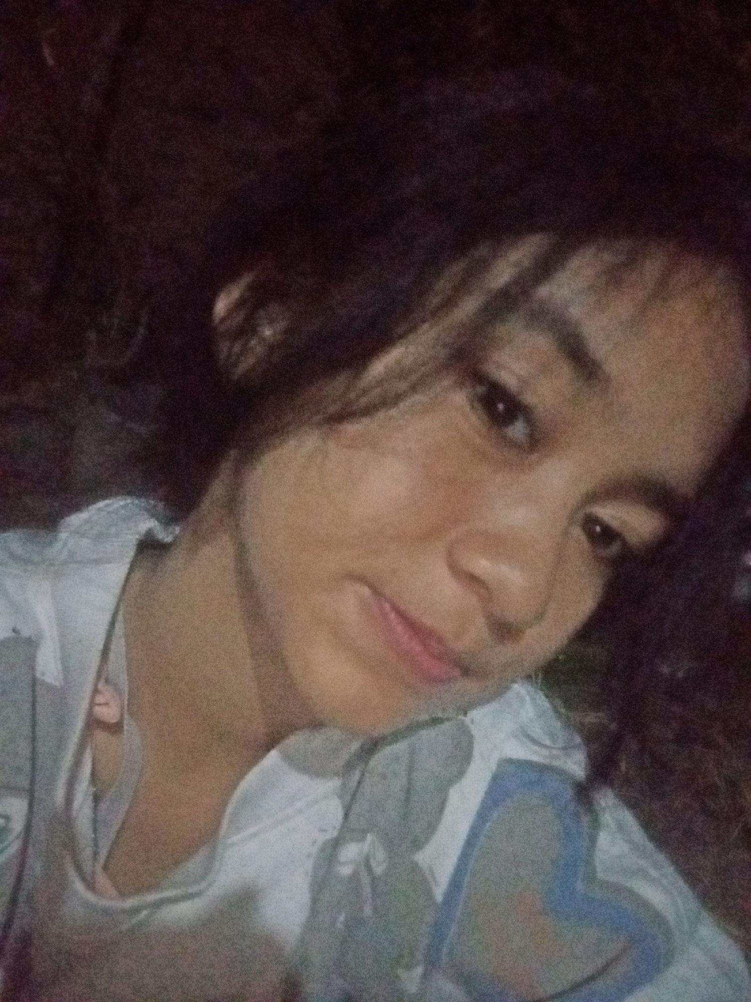 In a dimly lit, indoor setting, a young female of Southeast Asian descent is captured in a close-up, selfie-style photograph. She has dark, wavy hair framing her face and appears to be wearing a light-colored shirt with a pattern. The background is dark and indistinct, offering no clear clues about her surroundings within Badas, Indonesia. The lighting is low, creating a grainy texture across the image and obscuring finer details. Her facial expression is neutral, with her gaze directed slightly away from the camera.