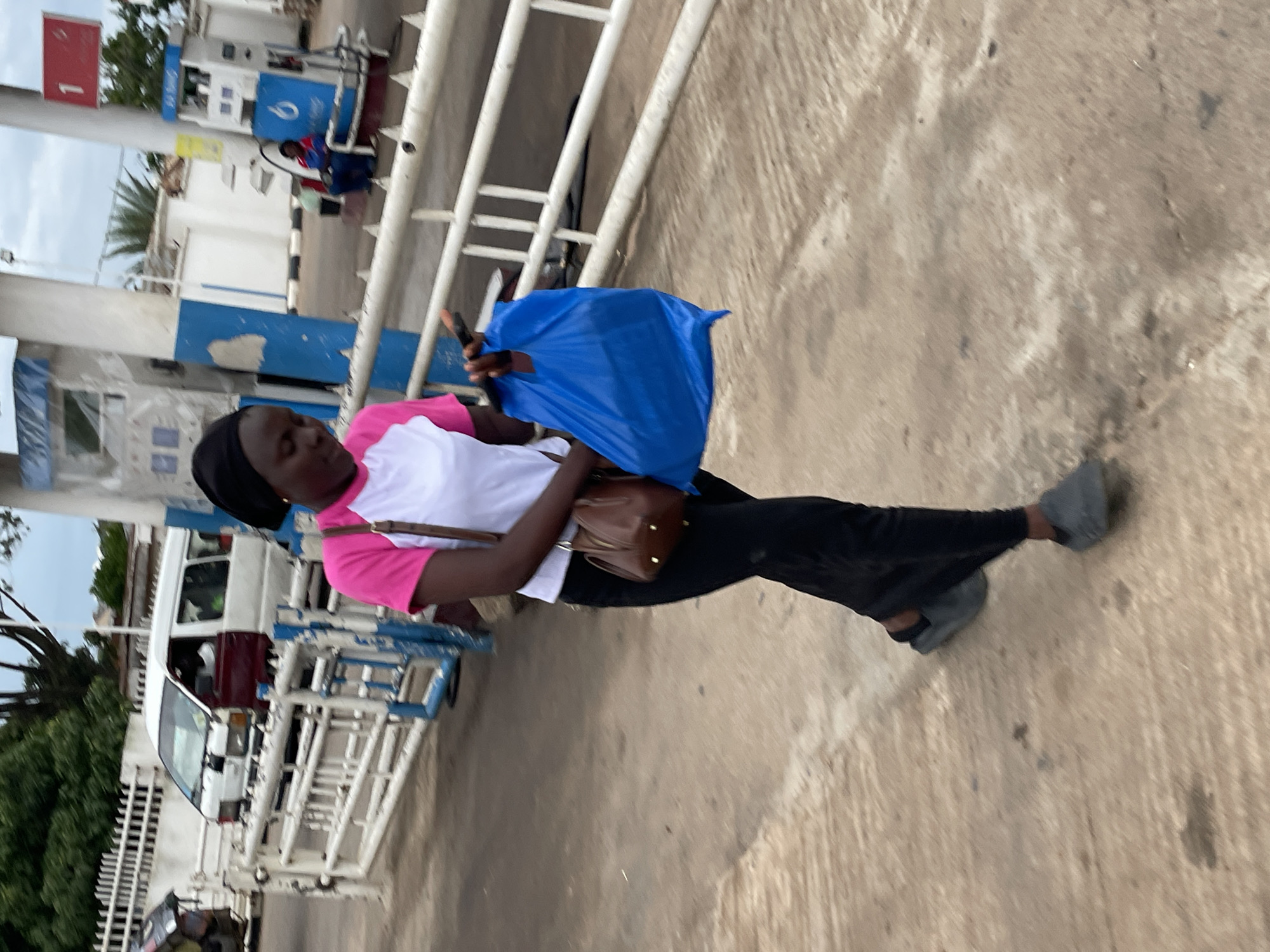 A young Black woman wearing a black headscarf and a white and pink shirt is walking past a gas station. She is carrying a blue plastic bag in her right hand and a brown purse on her left shoulder. The gas station has several pumps and is located on a paved road. In the background, a white van and a white fence are visible. The image appears to have been taken in Jos, Nigeria.