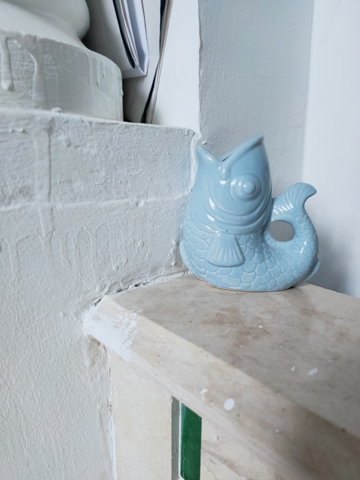 A light blue ceramic fish-shaped decorative object sits on a light beige ledge. The fish is oriented to the right, with its mouth open and a circular hole on its tail fin. The ledge is a part of a textured, cream-colored surface that extends upwards and to the left. To the left of the ledge, a white, textured wall with dripping paint is visible. Behind the fish and on the ledge, other objects are partially visible, including what appears to be a stack of books or papers. The overall environment suggests an indoor setting, possibly a shelf or windowsill. The specific location in Mrezga, Tunisia cannot be determined from the visual information alone.
