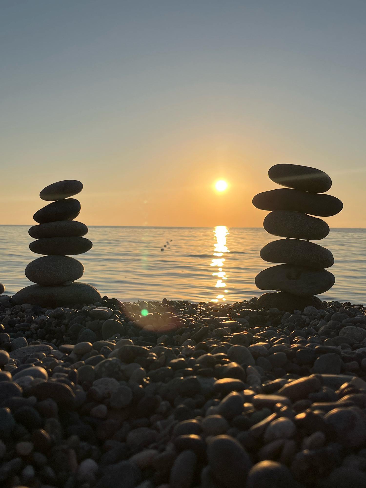 The image depicts a serene sunset scene on a pebble beach in Kobuleti, Georgia. Two towers of balanced stones stand prominently in the foreground, silhouetted against the warm glow of the setting sun. The sun is a bright orb positioned near the horizon, casting a shimmering reflection on the gentle waves of the sea. The sky transitions from a pale blue at the top to soft orange and yellow hues near the sun. The beach is covered in a variety of smooth pebbles, creating a textured foreground that leads the eye towards the water and the sky. The overall atmosphere is peaceful and tranquil.