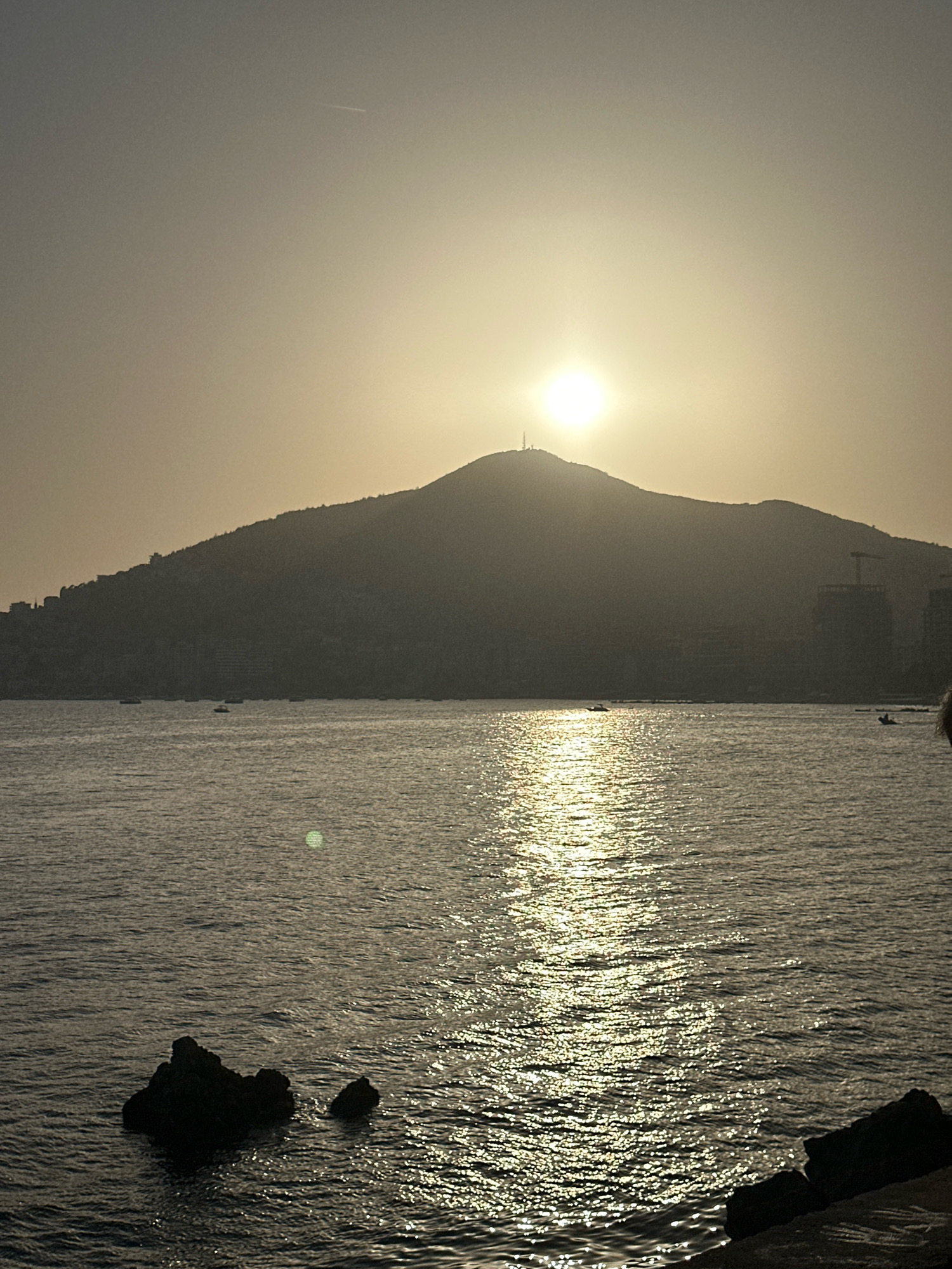 The image depicts a sunset over the sea, with a prominent hill silhouetted against the bright sky. The hill has buildings and a tower on its summit, suggesting it is an urban or semi-urban area. The water reflects the sunlight, creating a shimmering effect. A few boats are visible on the water, and rocks are present in the foreground. The overall scene is tranquil and atmospheric, characteristic of a coastal sunset. The presence of the hill with buildings and a tower is consistent with a view of Budva, Montenegro, where the iconic hilltop fortress and surrounding urban development are visible from the sea.