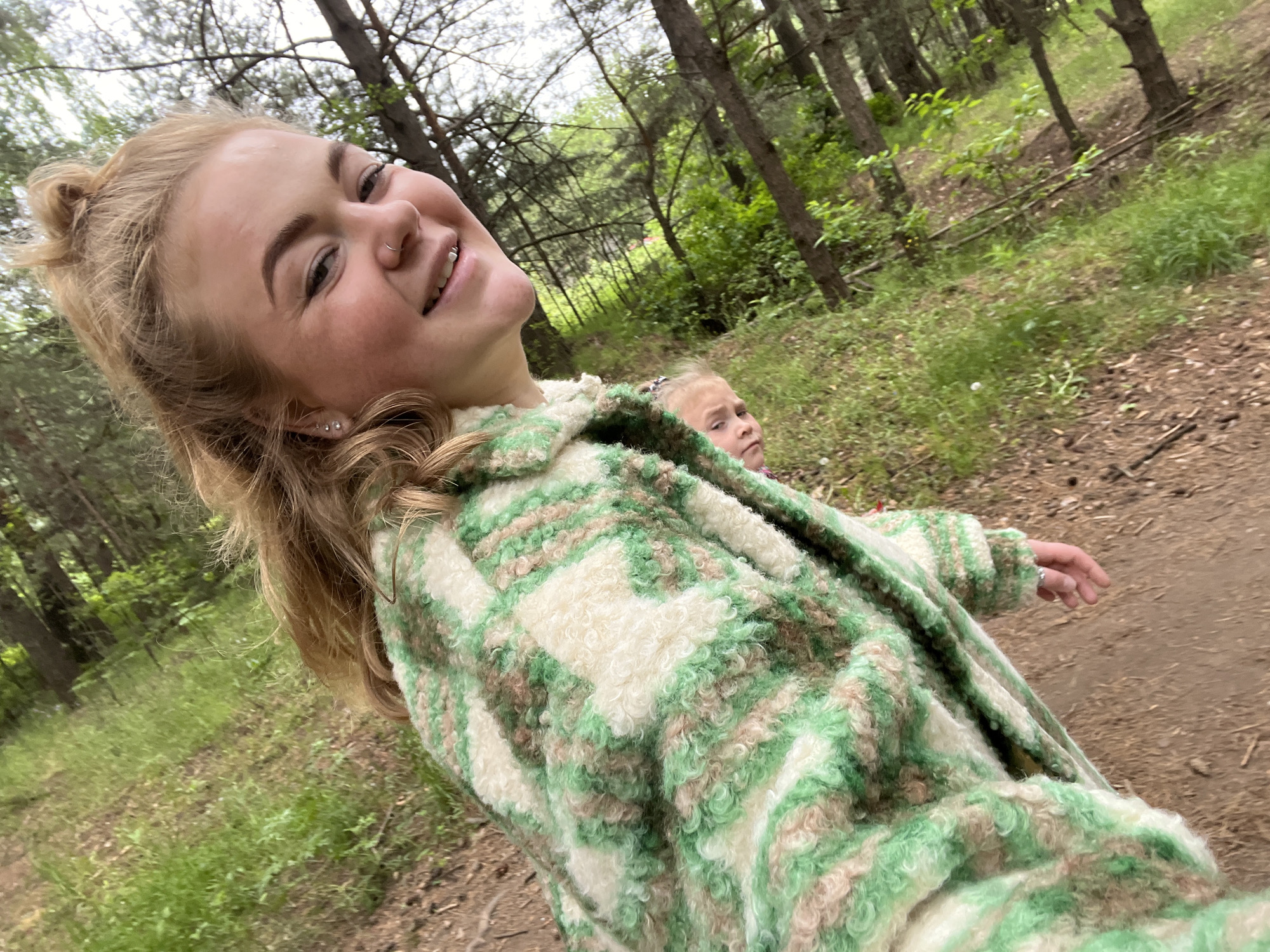 An outdoor, slightly tilted, selfie-style photograph captures a young woman and a child on a dirt path in a wooded area in the city of Chaikovsky, Russia. The young woman, occupying the left foreground, has blonde hair styled with a top bun, light skin, and is smiling broadly towards the camera. She wears two silver stud earrings, a silver nose ring, and a labret piercing. Her attire is a fuzzy, green, white, and light brown plaid jacket. Her right hand, visible at her side, displays a ring on the ring finger. A small child, with light hair, is partially visible behind and below the woman, looking towards the camera with a neutral expression. The path, composed of dirt and sparse vegetation, extends into the background, flanked by green grass and numerous trees with slender brown trunks and green foliage, indicating a forest or park setting. The lighting is natural and diffuse.