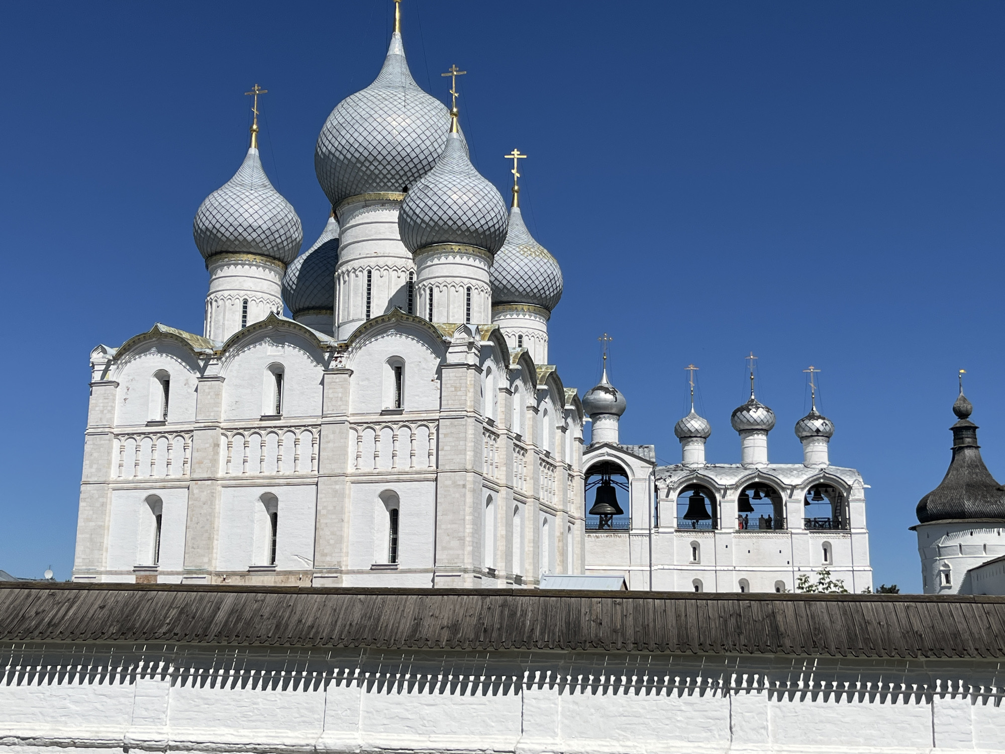 The image shows the Rostov Kremlin, located in the city of Rostov, Yaroslavl Oblast, Russia. The Kremlin is a large, white stone complex with several onion-domed churches and a bell tower. The domes are silver, and the crosses on top are gold. In the foreground, there is a white brick wall with a wooden roof. The sky is a clear blue. The image is taken from a low angle, looking up at the Kremlin. There are a few people visible on the bell tower.