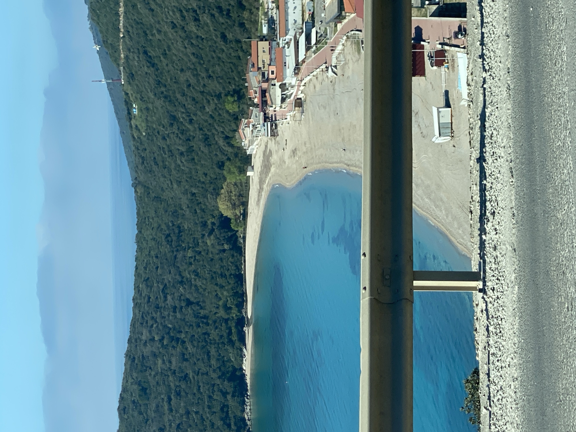 The image shows a coastal scene in Prijevor, Montenegro. A narrow beach borders a clear blue body of water. On the beach, several buildings line the shoreline. Dense green vegetation covers the hill adjacent to the beach. A gray metal support structure bisects the view, with a gray concrete road running parallel to the beach on the right side of the support. In the distance, another land mass is visible through a haze.
