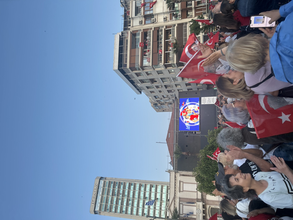 A large crowd of people are gathered in the Konak Square of İzmir, Türkiye, cheering and waving Turkish flags. A large screen displays an image of four people, two adults and two children, holding soccer balls. The people in the crowd are mostly young adults and middle-aged adults. Some children are also present. The crowd is standing on the street in front of a large building with a digital billboard. The sky is clear and blue. A tall building with the sign "EGE PALAS" is visible in the background. The image was captured in Konak Square, Izmir, Turkey at 38.437 latitude and 27.141 longitude. The crowd is facing the screen, and some are waving Turkish flags. The people in the crowd are all smiling and laughing, and the atmosphere is festive. Some of the people are wearing hats or scarves, and others are wearing casual clothing. The image is taken from a slightly elevated angle, giving the viewer a good perspective of the crowd and the surrounding buildings. The image is clear and well-lit.