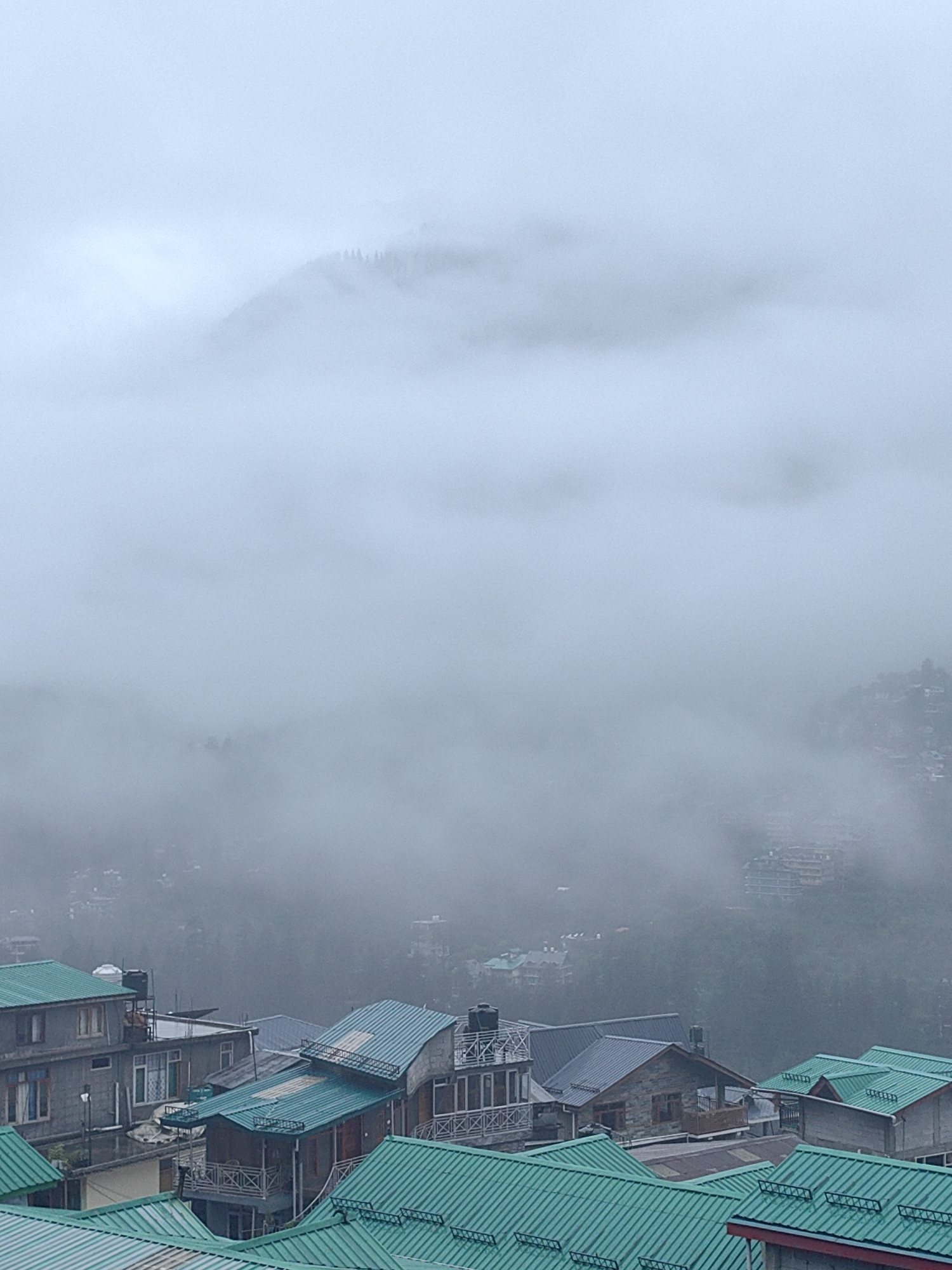 This is an aerial, overcast, medium-shot photograph of a mountainous village in Vashisht, India. The image is dominated by a heavy, grey mist that obscures much of the landscape. In the foreground, several buildings with distinctive, brightly colored teal roofs are visible. These structures appear to be houses or guesthouses, with multiple levels and visible windows and balconies. Some buildings have exposed stone facades, while others are constructed from concrete. Water tanks are visible on the roofs of some structures. In the middle ground and background, the mist thins slightly to reveal more buildings nestled into the hillside, also covered in mist. The distant mountains are almost entirely hidden by the thick fog, with only the faint outline of a forested peak visible at the very top of the frame. The atmosphere is subdued and hazy due to the weather conditions. No people or animals are discernible in the image.
