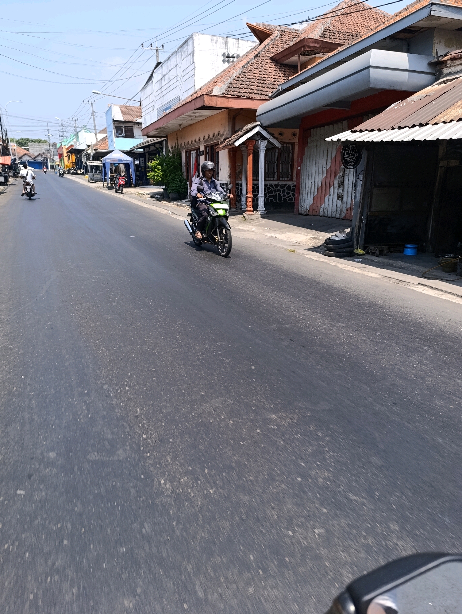 The image depicts a street scene in Glagah Sari, Indonesia, under a clear blue sky. The street is paved with asphalt and appears to be a typical urban road with buildings lining both sides. On the right side of the road, there is a shop with a corrugated metal roof, and stacked tires are visible outside. Further down the street, a motorcycle rider, wearing a helmet and a dark shirt with long sleeves, is moving away from the camera. The motorcycle is green and black. Other motorcycles and some people are visible in the background, further down the street, on the left side. The buildings on the left side of the street are varied in color and architectural style, with some appearing to be shops or small businesses. Power lines are strung overhead. The overall impression is of a busy, everyday street in a town or city in Indonesia.