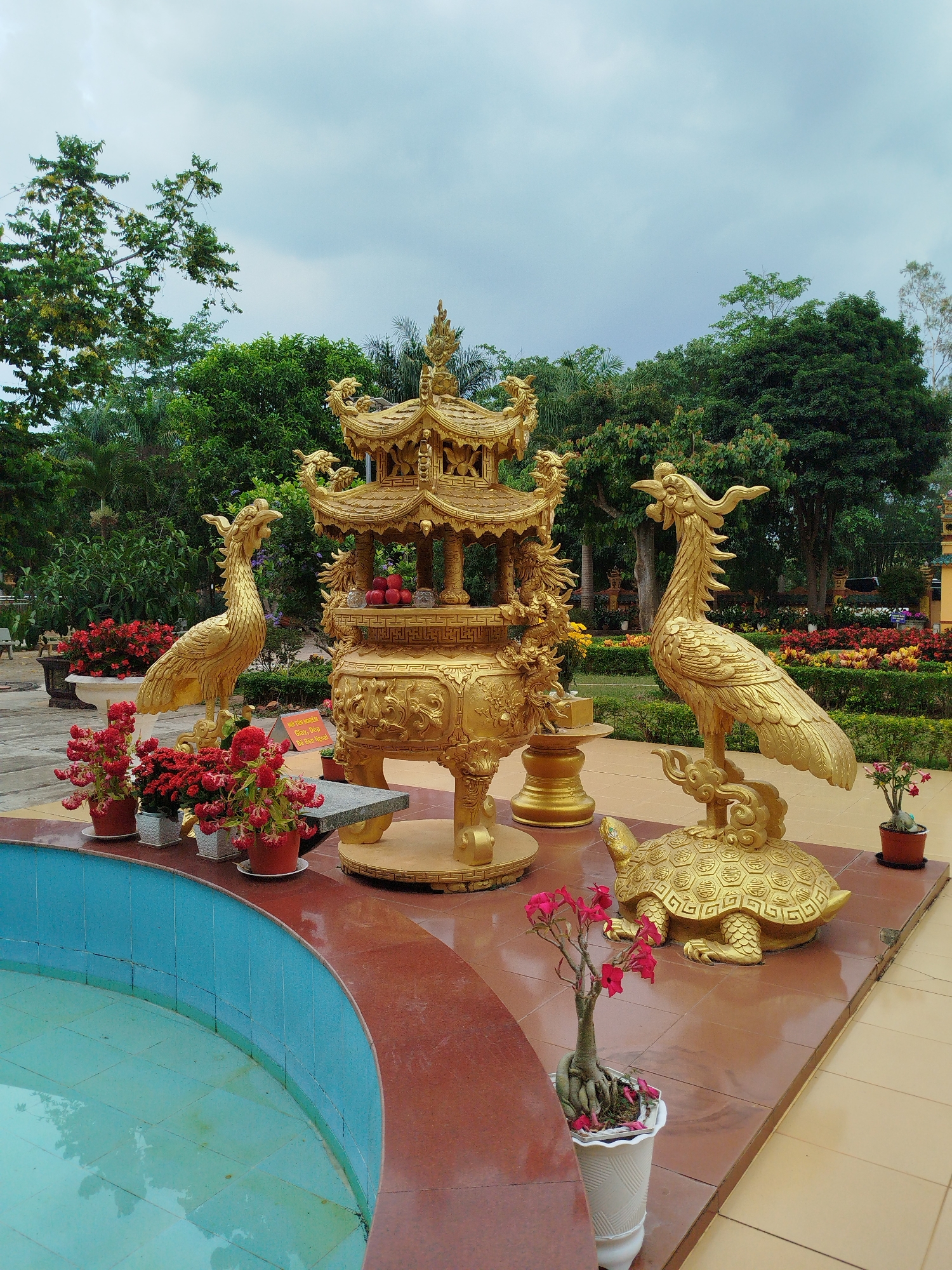 Captured at the temple in Phú An, Vietnam, the image features a golden, ornately decorated incense burner at the center of the frame, adorned with dragons and topped with a miniature pagoda-like structure. To its left, a golden phoenix statue stands amidst red potted flowers along a curved pool with blue tiles and a red rim. To the right, another golden phoenix statue is perched on a golden turtle. The background includes lush greenery, trees, and a cloudy sky. The ground is covered with red and yellow paving stones. A small sign is placed next to the incense burner.