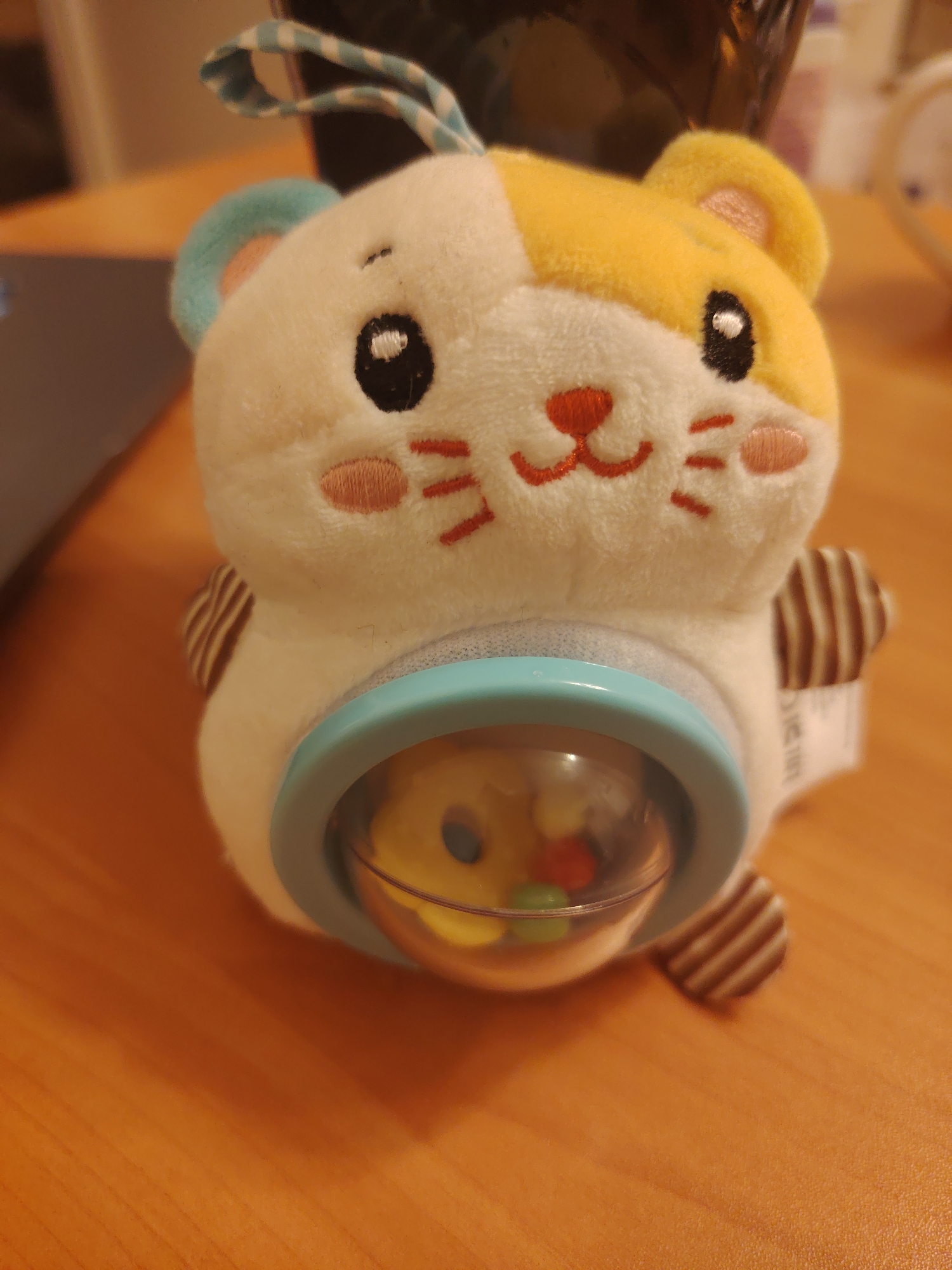 A close-up, eye-level shot shows a stuffed toy on a wooden table. The toy is shaped like a hamster and is white with yellow markings on its head. It has black eyes, a red nose and mouth, and pink cheeks. There is a clear plastic sphere on the front of the toy, which contains colorful beads. The toy has striped brown ears and a blue loop on top. The background is blurred, but appears to be a room with a laptop and a dark object. There is no indication of the city of Konakovo, Russia.