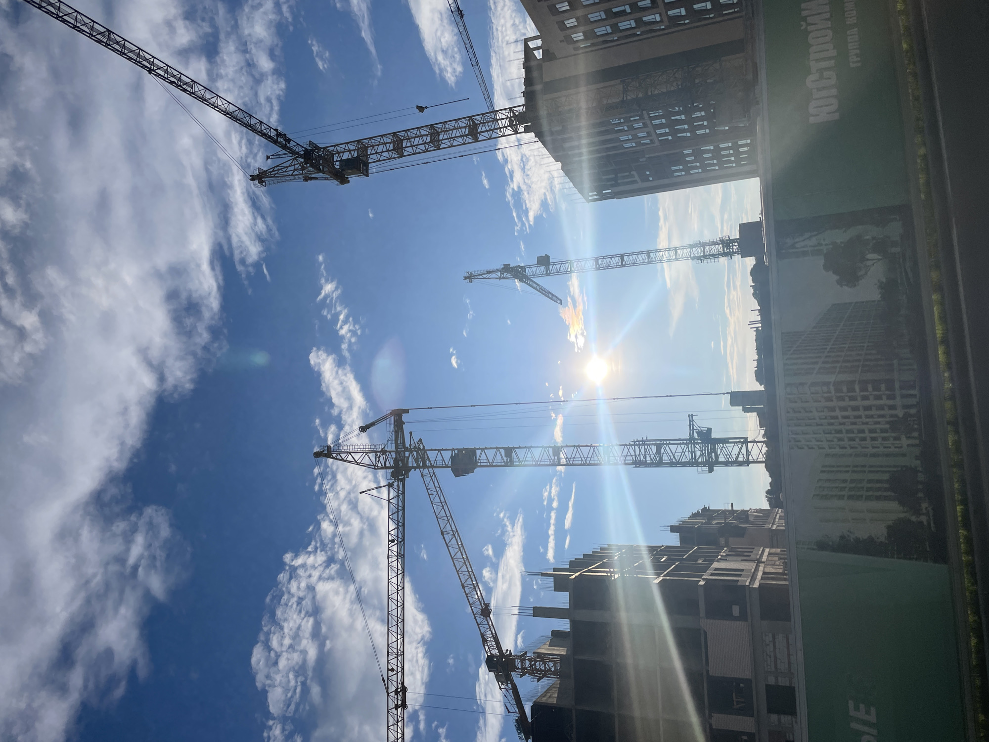 The image shows a construction site with multiple tower cranes silhouetted against a bright blue sky with scattered clouds. The sun is visible, creating lens flares and sunbeams that cut through the scene. In the foreground, there are partially constructed buildings with visible rebar and concrete structures. Further back, completed buildings with windows are also visible. A green construction fence with Russian text and images of buildings is in the lower portion of the frame. The construction site is located in Stavropol, Russia.