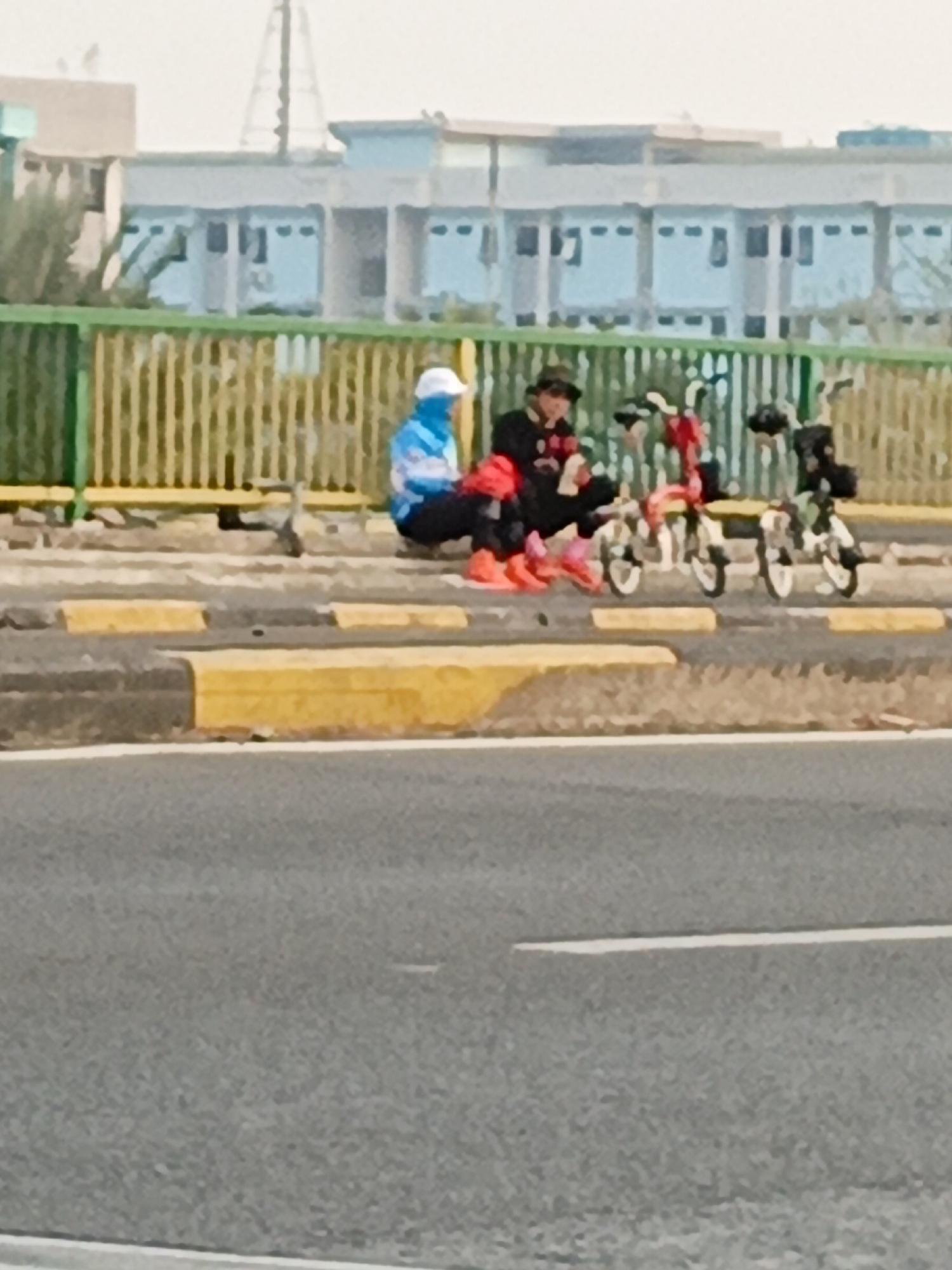 Two individuals are seated on a curb next to a yellow and green railing, with three foldable bicycles partially visible beside them. One person, wearing a blue and white hooded jacket and a white hat, is seated facing the right, with their legs bent. The other person, wearing a dark jacket and a hat, is seated facing the left, with their legs crossed. The background shows a light-colored building with blue windows. The scene appears to be located on a street in East Jakarta, Indonesia.