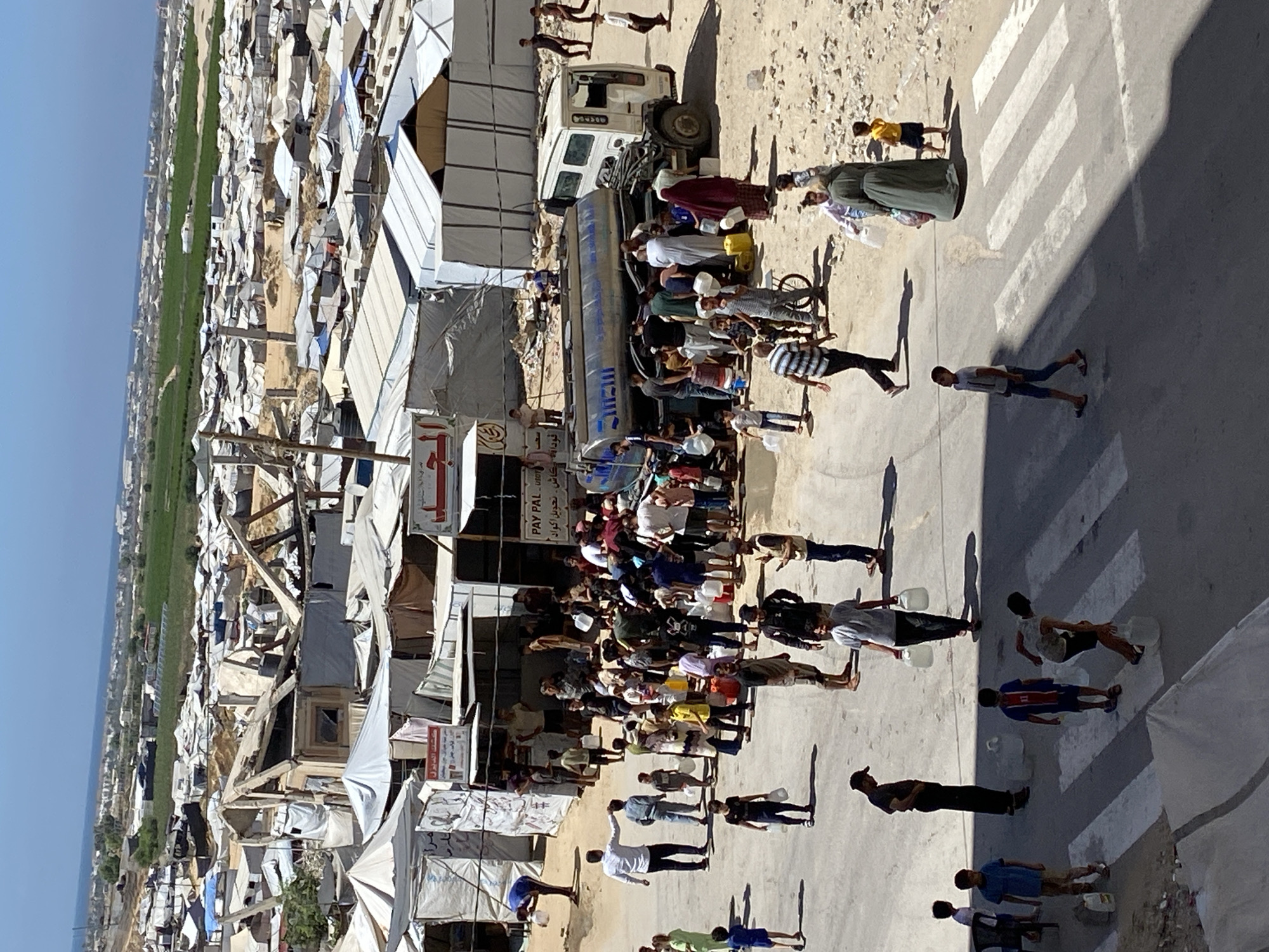 The image depicts a bustling scene in Khan Yunis, Palestinian Territories, where a water tanker truck is distributing water to a crowd of people. The environment appears to be a makeshift settlement, with numerous tents and partially constructed shelters scattered across a sandy terrain. The sky is clear and blue, indicating a sunny day.
In the foreground, a group of individuals, including men, women, and children, are gathered around the water tanker. Many are holding plastic containers and buckets, actively collecting water. Some are waiting in line, while others are in the process of filling their containers. The expressions on their faces suggest a sense of urgency and necessity.
The water tanker truck is a prominent feature, with its large tank visible. The truck appears to be parked on a road or pathway within the settlement.
In the background, more tents and shelters stretch out as far as the eye can see, forming a dense settlement. Some structures show signs of damage or disrepair, hinting at potential conflict or harsh living conditions. Beyond the settlement, a wider landscape is visible, with some greenery and buildings in the distance.
The overall impression is one of a community striving to meet its basic needs amidst challenging circumstances. The image captures a moment of collective effort and resilience, as people come together to secure essential resources.