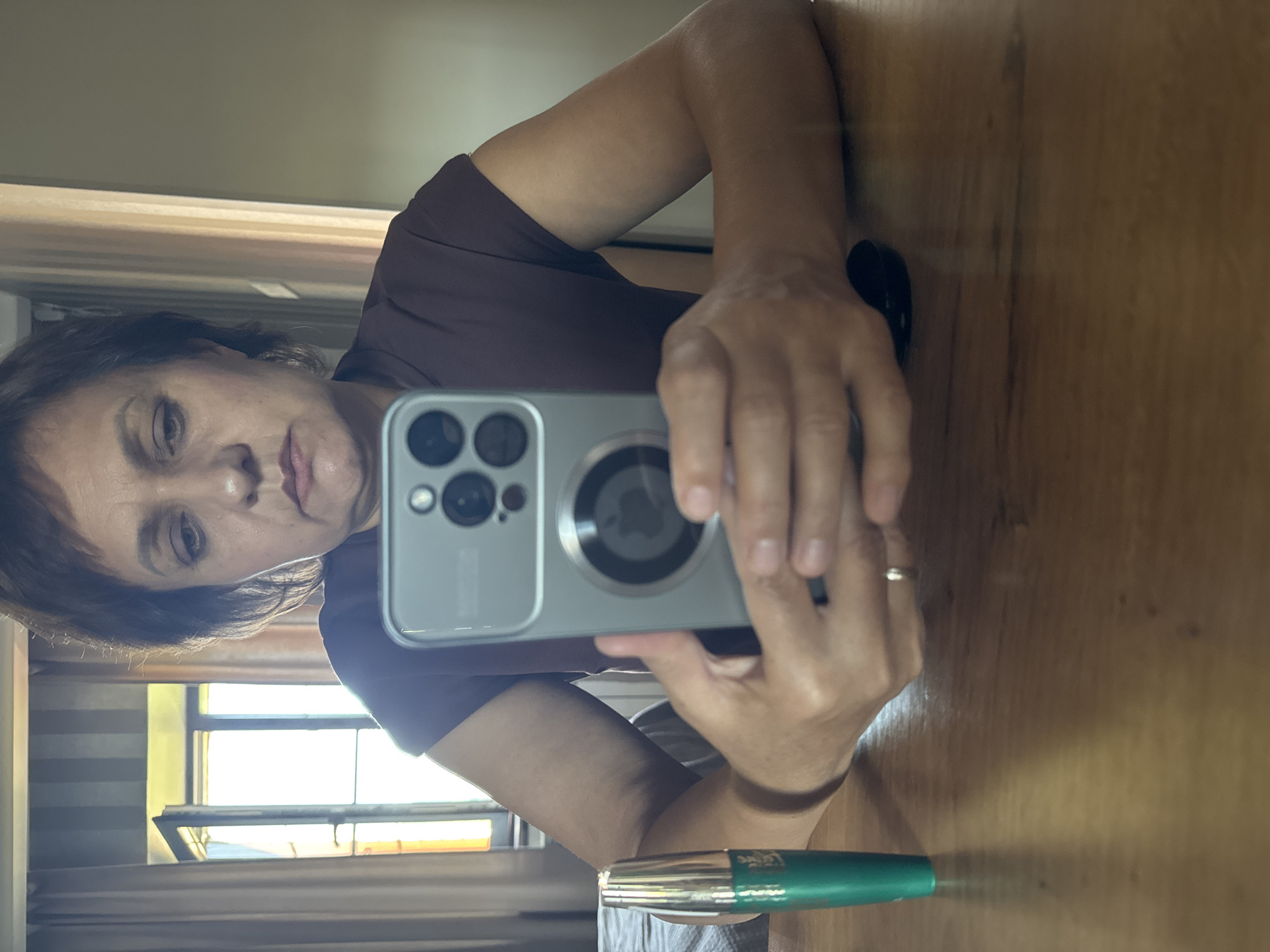A woman with short, dark brown hair sits at a wooden table, holding a smartphone in her hands and looking at its screen. She is wearing a dark brown, short-sleeved shirt. A green tube, likely mascara, stands upright on the table to her left. In the background, a window is visible behind drawn curtains, and the reflection of the woman and the room can be seen in the phone's screen and possibly a mirror. The scene appears to be indoors, possibly in a home, with natural light entering from the window. The location is identified as Soloneshnoye, Russia.