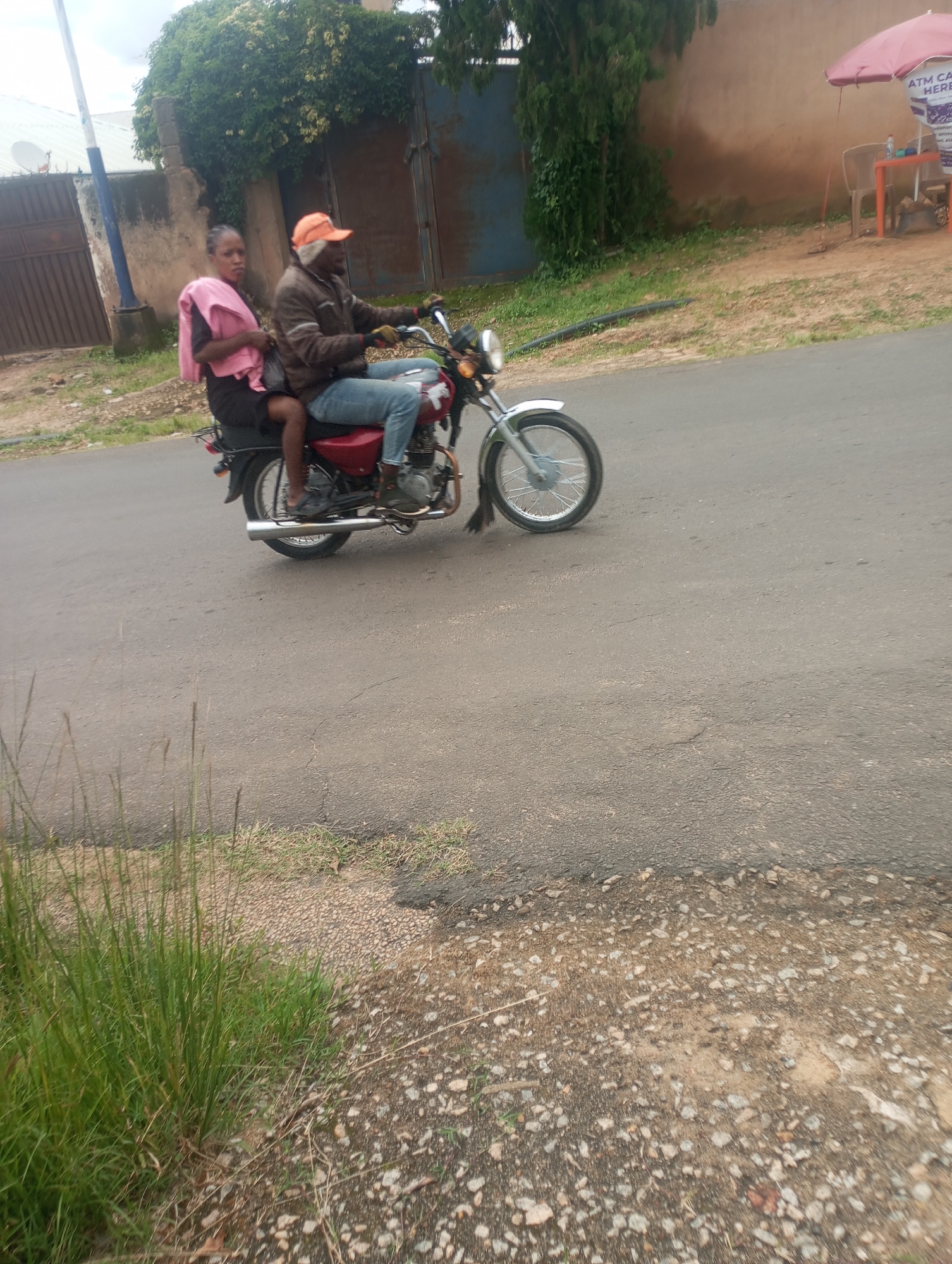 An adult male, wearing an orange cap, a brown jacket, and blue jeans, rides a red motorcycle on a paved road. He is seated and operating the vehicle. A young female, wearing a pink headwrap or scarf and a black skirt, sits behind him, holding onto him. The motorcycle has a silver exhaust pipe and a silver fender over the front wheel. The scene appears to be in Jos, Nigeria, based on the style of the buildings and vegetation. In the background, there is a large blue metal gate and a building with a cream-colored wall. To the right, a pink umbrella shades a small table and chairs, with a sign indicating "ATM CASH HERE." The road surface is asphalt, with a gravelly shoulder and dry grass on the side. The sky is overcast.