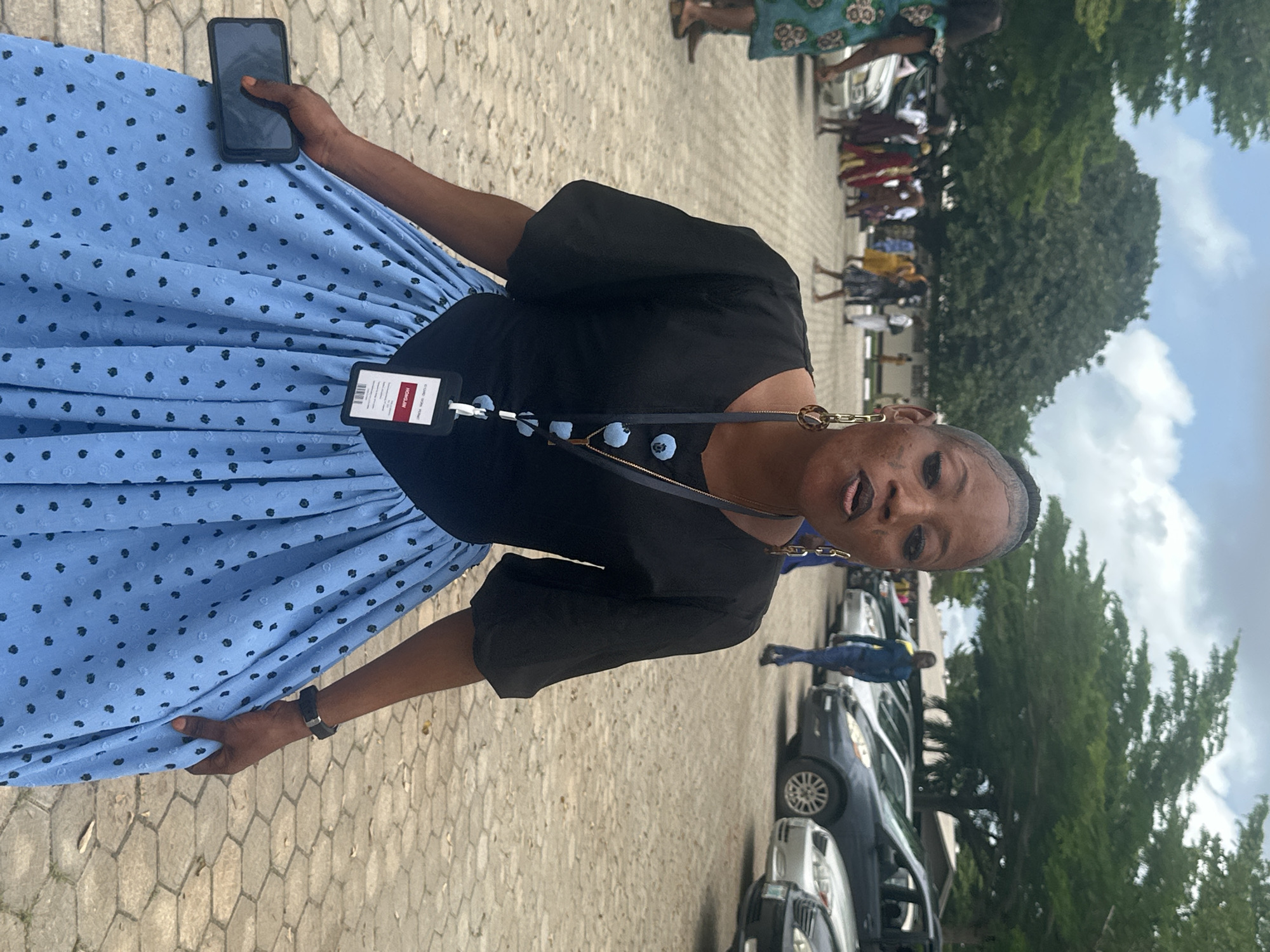 A woman is standing in a parking lot in Itoga, Nigeria. She is wearing a black top with puff sleeves and a light blue skirt with black polka dots. She has on large hoop earrings and a necklace with a pendant. A lanyard with an ID badge is hanging around her neck. She is holding a dark-colored smartphone in her left hand. The parking lot is paved with interlocking stones. In the background, there are several cars parked, and people are visible in the distance, some walking and others standing. Trees line the edge of the parking lot, and the sky is cloudy.
