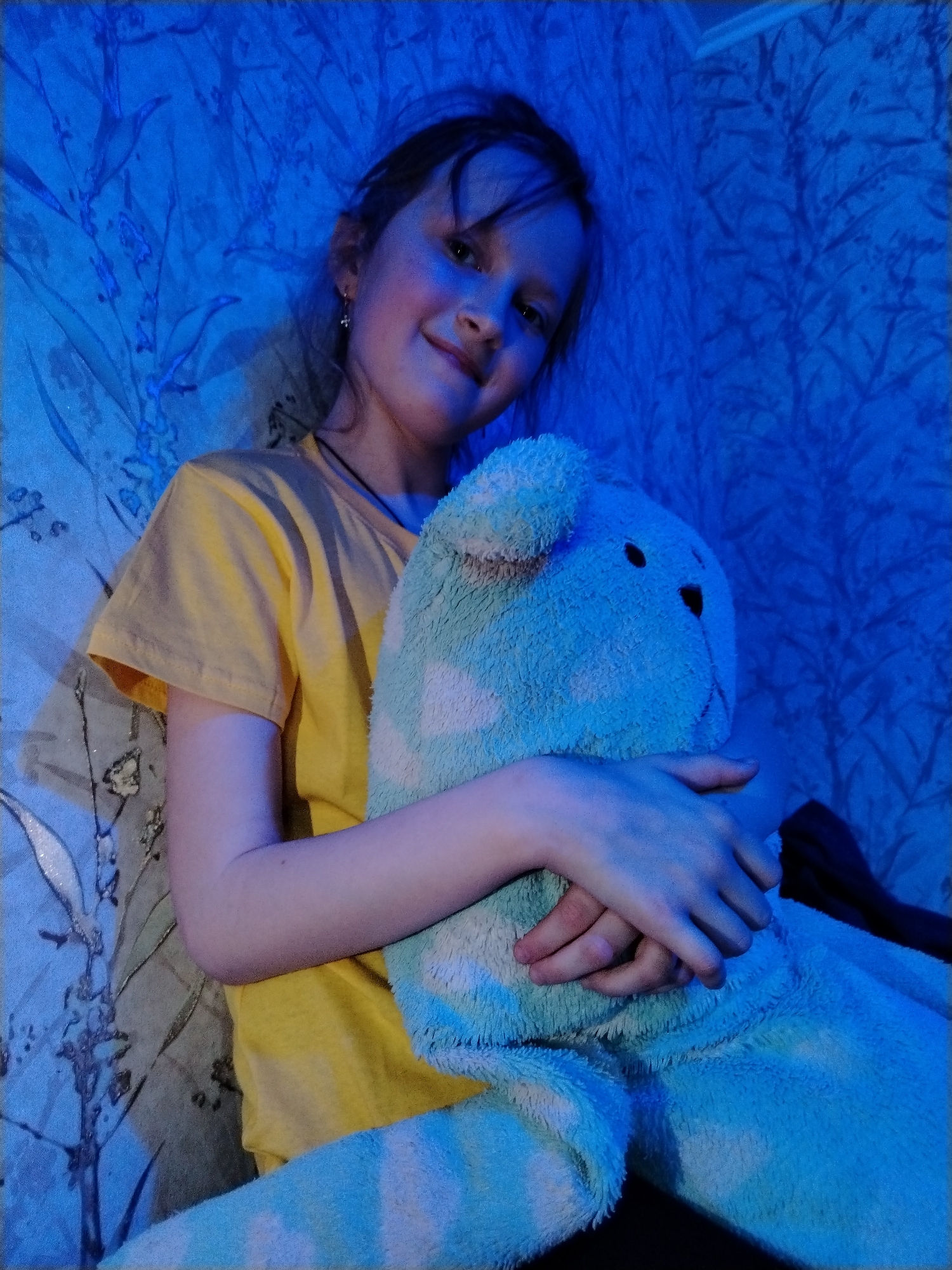 In an indoor setting within Nizhnyaya Gar', Russia, a young female, appearing to be a child or pre-teen, is seated and directly facing the camera. She has dark hair with bangs and fair skin, exhibiting a subtle smile. She is dressed in a yellow short-sleeved t-shirt and a dark choker-style necklace, with a small cross-shaped earring visible in her left ear. Her arms are wrapped around a large, light blue plush stuffed animal, which features white patches and has dark, round eyes visible on its head; her hands are clasped over the animal's torso. The background is a wall covered in wallpaper, displaying a light blue floral or vine pattern, possibly with reflective elements, against a darker base. The entire scene is distinctly illuminated by a pervasive blue light source.