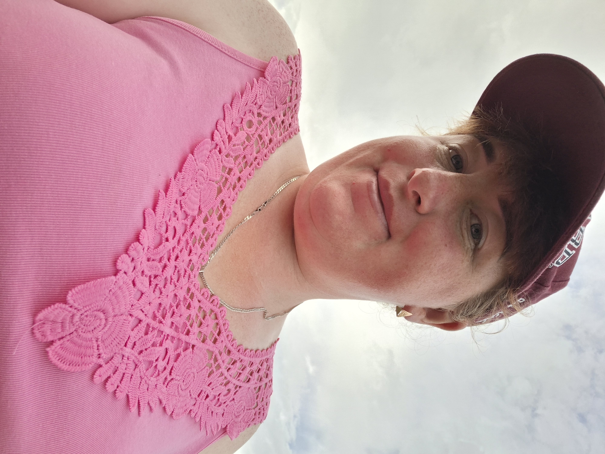 A close-up, low-angle, outdoor selfie of a woman wearing a maroon baseball cap and a pink tank top with lace trim. She has fair skin with freckles and is looking directly at the camera with a slight smile. The background is a cloudy sky. A gold, triangular earring is visible in her right ear, and a thin silver chain necklace is worn around her neck. The woman appears to be in her 30s or 40s, and her hair is reddish-brown, peeking out from under the cap.