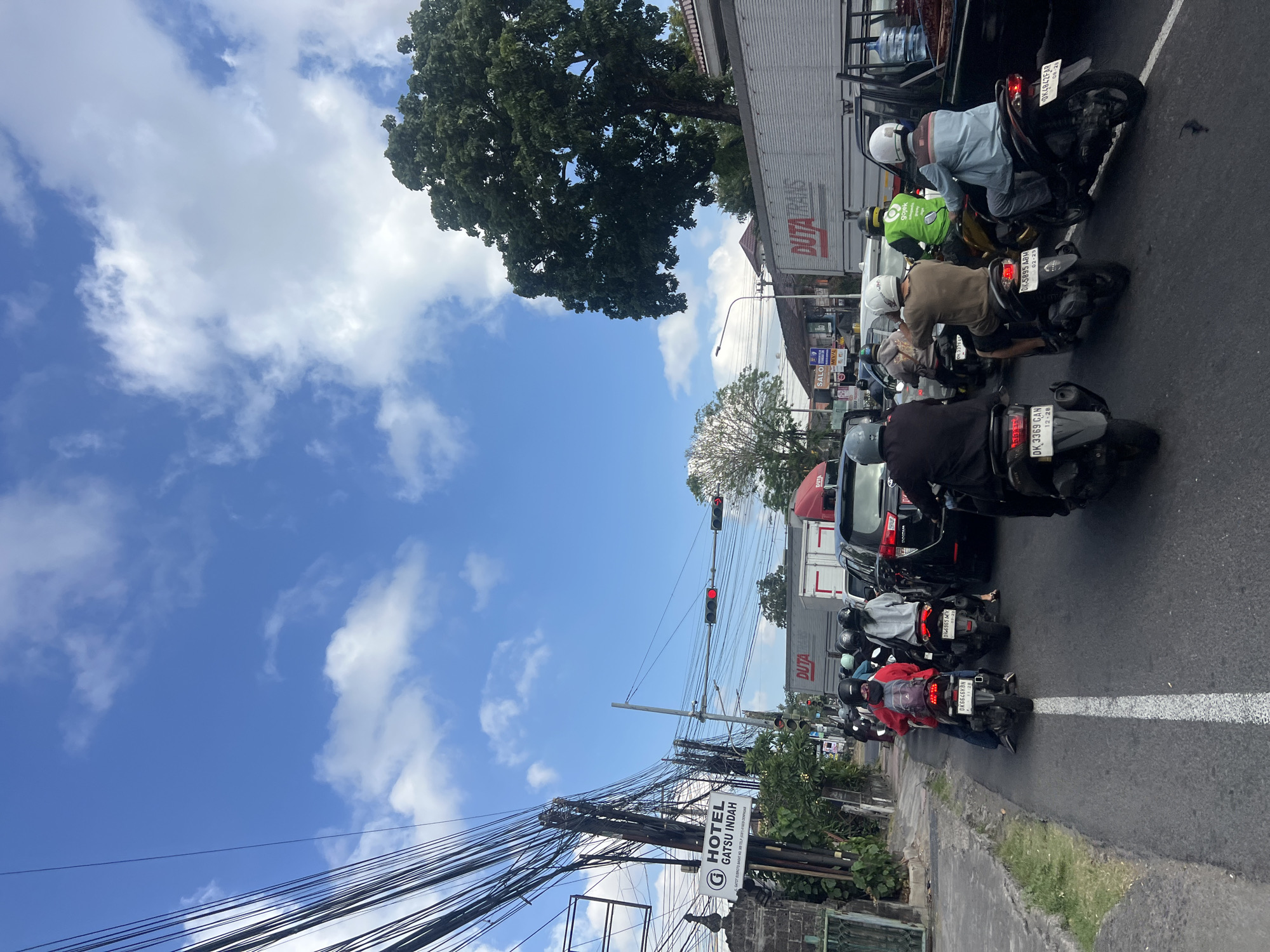 The image depicts a busy street scene in Denpasar, Indonesia, likely during daylight hours, under a bright blue sky with scattered white clouds. The foreground is dominated by a traffic jam of motorcycles and cars, indicating a congested urban environment. A prominent white line divides the road, with several motorcyclists lined up. Some riders are wearing helmets, and their clothing varies in color and style, suggesting everyday commuting.
On the left side of the road, a sign for "HOTEL GATSU INDAH" is visible, along with a weathered stone wall and lush green foliage, hinting at the presence of established buildings and vegetation. Overhead, a complex network of utility wires crisscrosses the sky, a common sight in many Indonesian cities.
Further into the scene, large trucks are present, including one with "DUTA TRANS" branding. The overall atmosphere suggests a typical day in a bustling Indonesian city, characterized by heavy traffic, a mix of vehicles, and the distinct visual elements of its urban landscape. The presence of a "Gojek" branded vest on one of the motorcyclists further confirms the Southeast Asian setting.