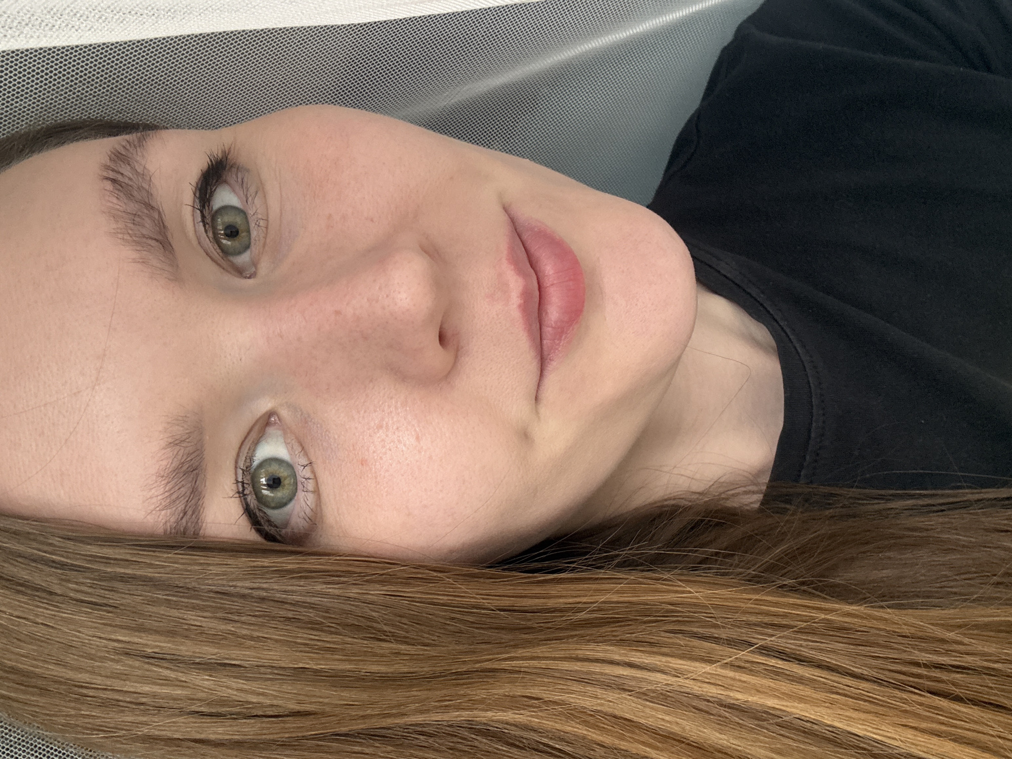 A close-up, angled portrait shows a light-skinned woman with light brown hair and green eyes lying on a surface with a white mesh covering. She is wearing a black shirt. The background is out of focus, suggesting an indoor setting. There is no indication of the location being Izhevsk, Russia. The image appears to be a personal photograph.