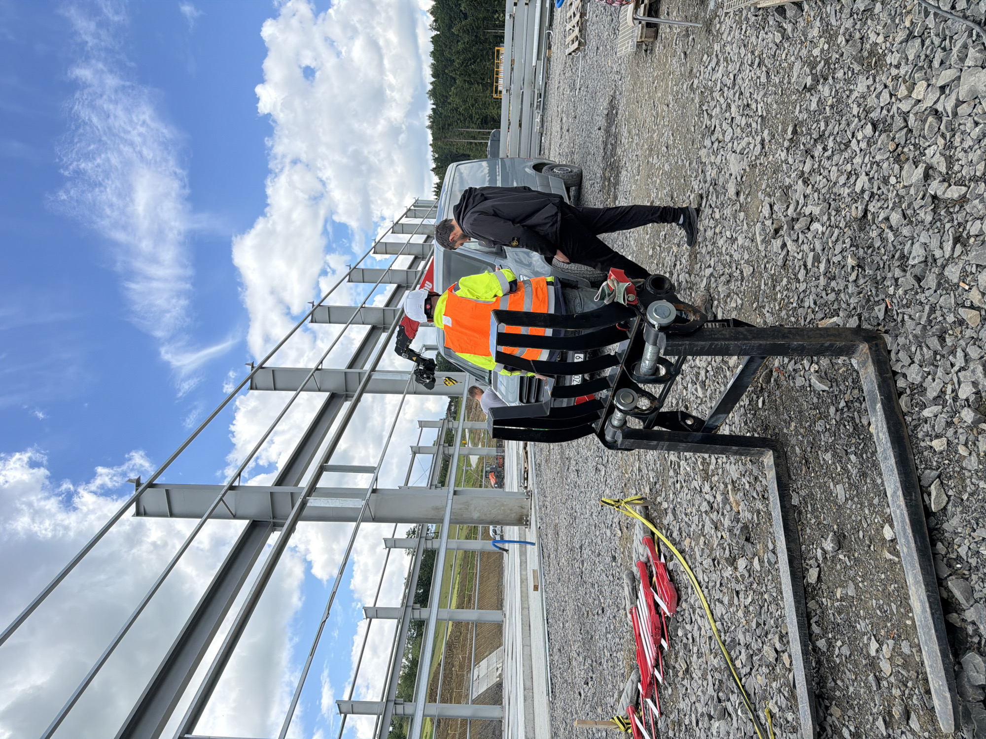 The image depicts an outdoor construction site in Stróża, Poland, with a large steel structure under construction. In the foreground, a forklift attachment with two large prongs rests on a gravel surface. Two men are visible in the midground. One man, wearing a bright orange high-visibility vest and a white hard hat, stands facing away from the camera. He appears to be operating a piece of equipment attached to a van. The second man, dressed in casual dark clothing, stands with his back to the camera, looking towards the first man and the van. A grey van is parked behind the forklift and near the men. In the background, the skeletal framework of a large building is taking shape, supported by concrete foundations and numerous steel beams. Beyond the construction area, a treeline marks the edge of the site, with a gently sloping, grassy hill visible to the left. The sky above is blue with scattered white clouds.