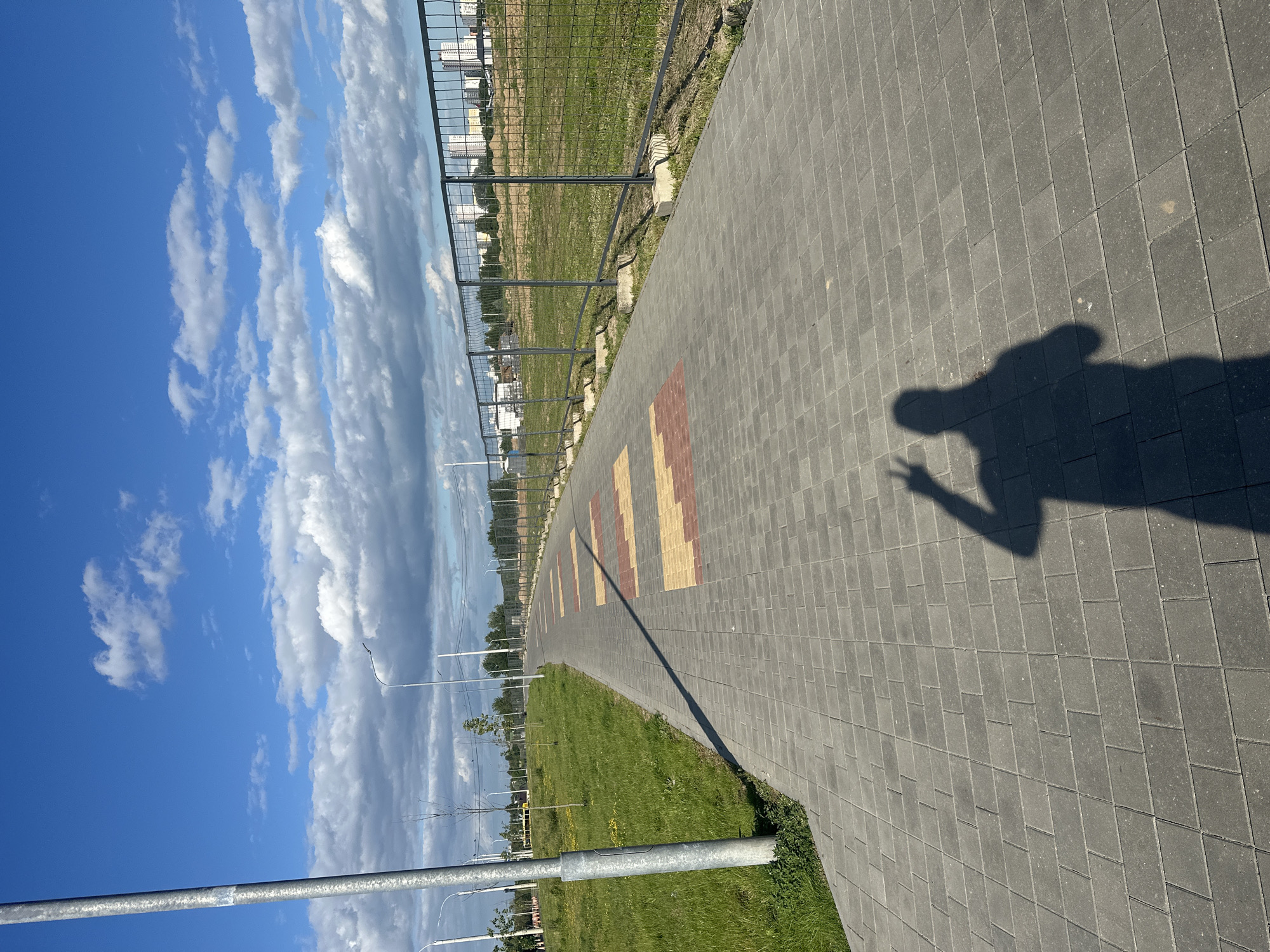 The image shows a paved walkway in a city park. The walkway is made of gray pavers and is lined with grass and trees. There is a fence on the right side of the walkway, and beyond the fence, there are buildings in the distance. The sky is blue with some clouds. In the foreground, there is a shadow of a person making a peace sign. The location is an urban area in Minsk, Belarus.