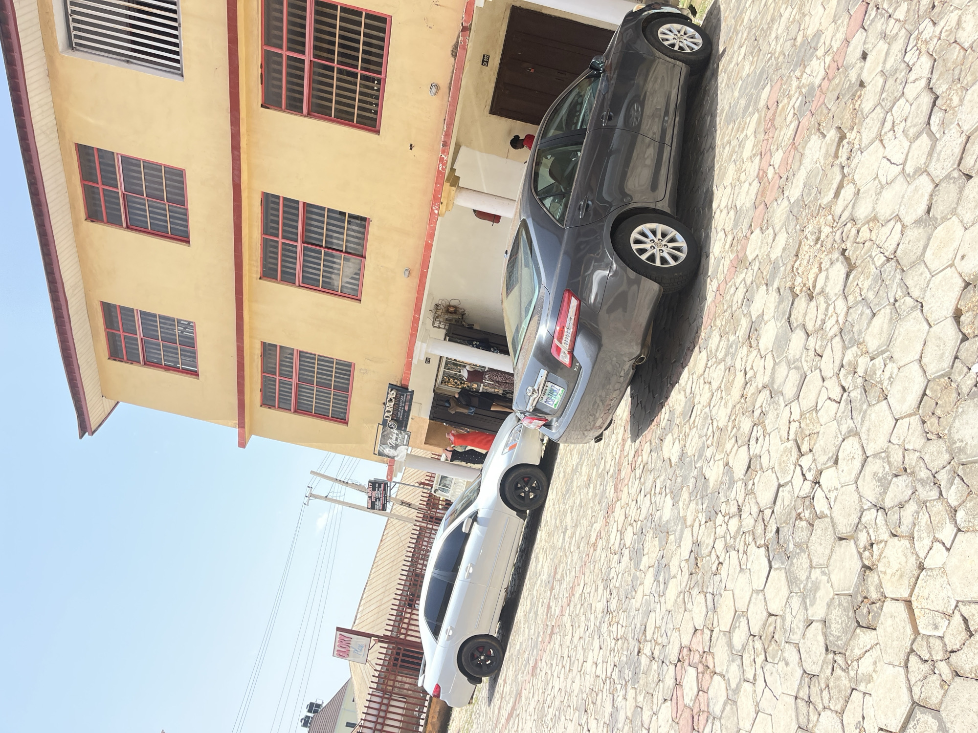 The image depicts a street scene in Gura Top, Nigeria, under a clear, sunny sky, suggesting daytime. The foreground is dominated by a paved walkway with a hexagonal pattern of light-colored stones. Two cars are parked on this walkway. The first car, a dark gray sedan, is positioned parallel to a building. The second car, a white sedan, is parked behind the first.
The building on the left is a two-story structure with a light yellow facade. It features several windows with red frames and metal grilles. The upper floor has a large, horizontally slatted vent. The lower floor has an entrance with white pillars and what appears to be a shop or business inside, with mannequins displaying clothing.
Several signs are visible in the image. One sign, partially obscured, has the text "DORCAS". Another sign, further down the street, reads "GLORY Place". There are also utility poles and wires. The overall impression is of a commercial or residential area on a bright, clear day. No specific activities or events are clearly depicted beyond the presence of parked cars. The scene appears ordinary and mundane, with no strong emotional cues.