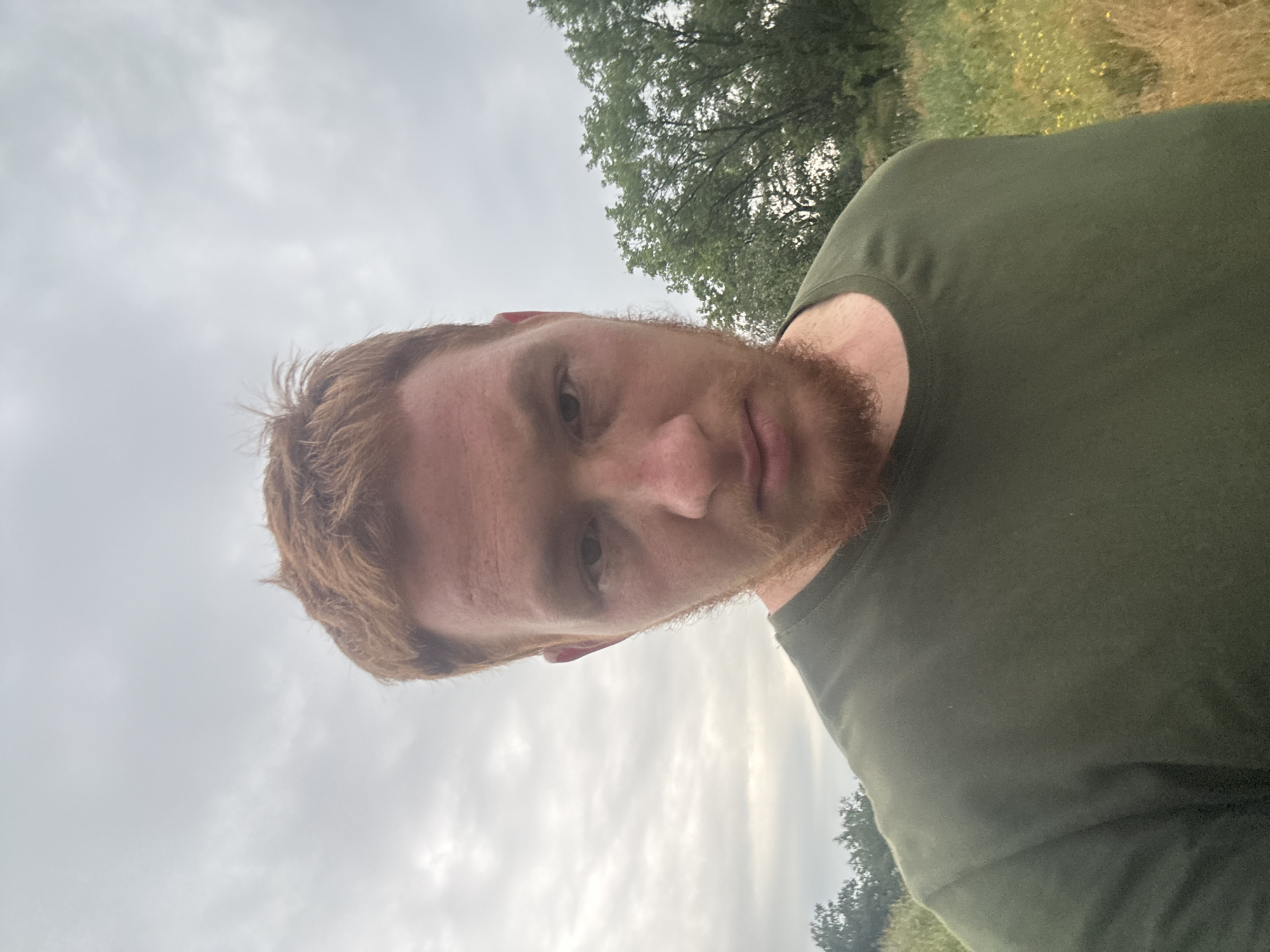 A medium shot photograph shows a person with red hair and beard facing the camera directly. The person is wearing a green t-shirt. The background features a cloudy sky, trees, and a field of grass with some yellow wildflowers. The image appears to be taken outdoors.