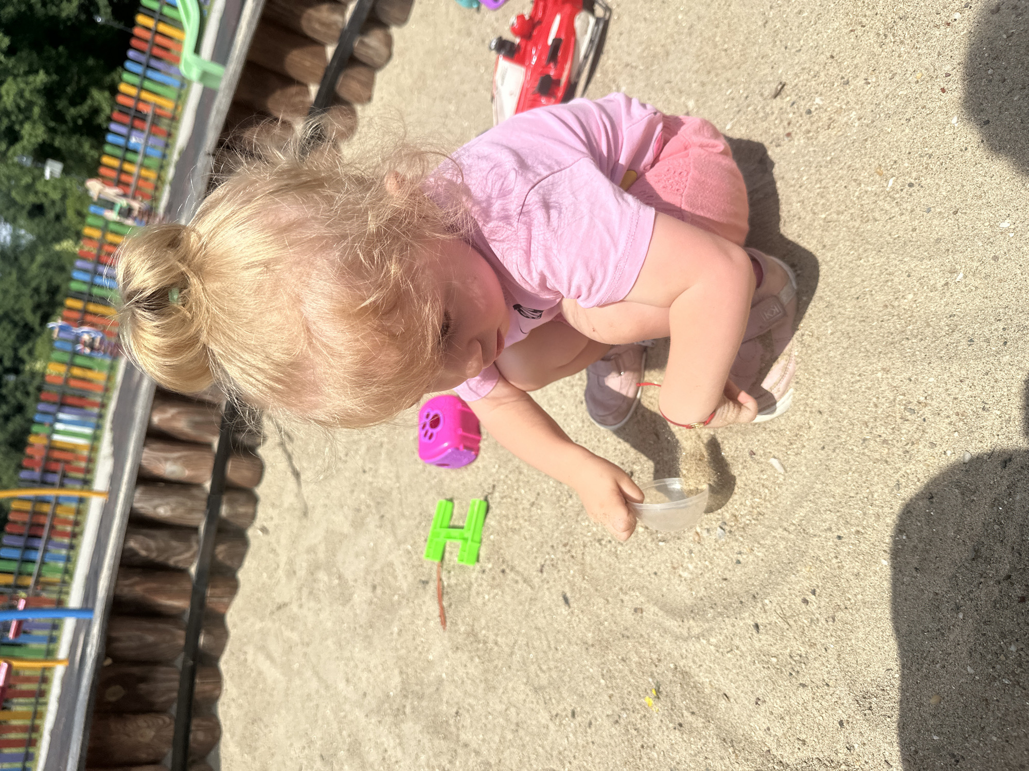 A young blonde girl with her hair in a bun is playing in a sandbox in Donchovci, Bulgaria. She is wearing a pink t-shirt and pink shorts, and is kneeling in the sand. The girl is holding a clear plastic cup and appears to be pouring sand from it. There are several children's toys scattered around the sandbox, including a green "H" shaped toy and a pink toy with holes. In the background, a colorful fence of a playground is visible, along with trees and foliage. The ground is covered in sand, with scattered pebbles and small shells. The overall lighting suggests a sunny day.