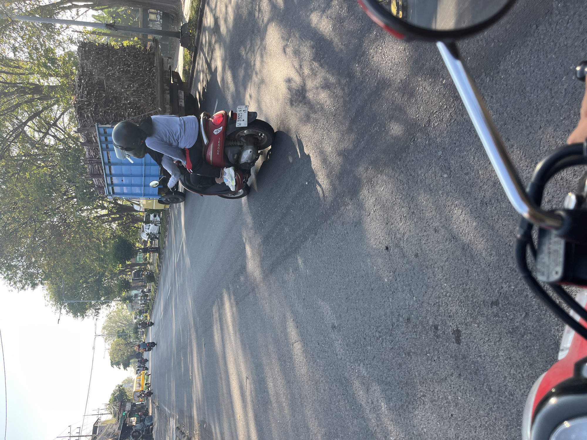 In Kludan, Indonesia, on a sunlit street, two individuals are riding a red scooter. The passenger, wearing a gray long-sleeved shirt and black pants, sits behind the driver, who is wearing a black helmet. In the background, a blue truck is loaded with what appears to be sugarcane stalks. The scooter's license plate reads "W 3925 NF". The scene is captured from the perspective of another vehicle, with a partial view of its handlebars and side mirror in the foreground. The road is asphalt and lined with trees casting shadows.