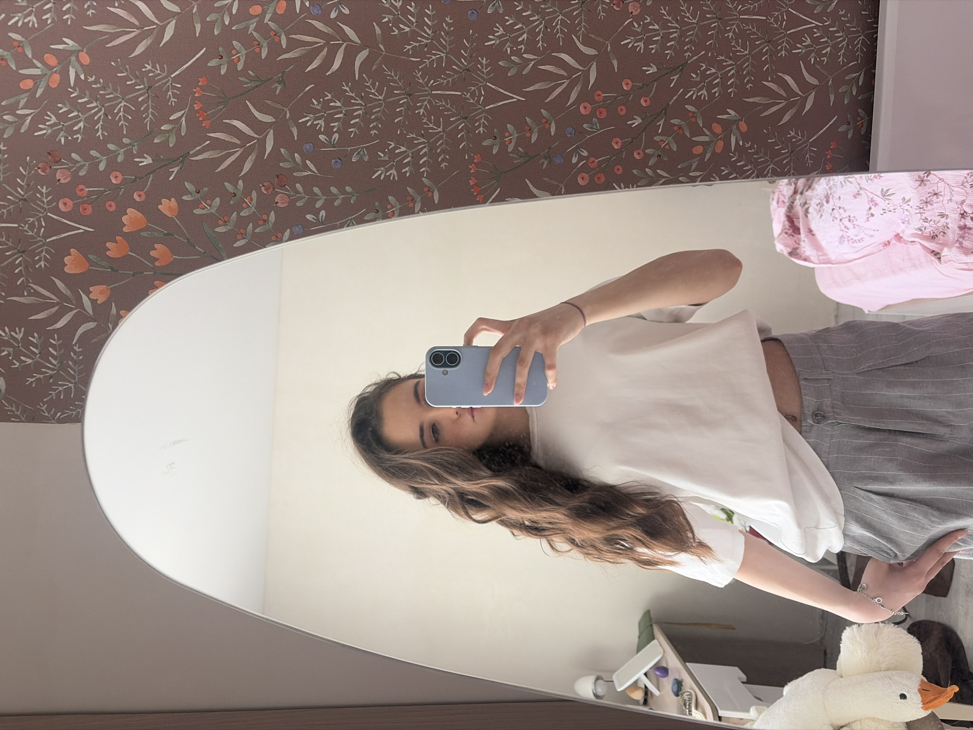 A young woman is reflected in a large, curved mirror against a backdrop of brown wallpaper with a floral pattern. She is holding a light blue smartphone in her right hand, presumably taking a selfie. The woman is wearing a white cropped t-shirt and grey striped pants. Her long, wavy brown hair falls over her shoulder. The reflection also shows a cluttered shelf with a plush white goose toy and other miscellaneous items. The setting appears to be indoors, likely a bedroom. The image lacks specific details to confirm it is in Izhevsk, Russia.