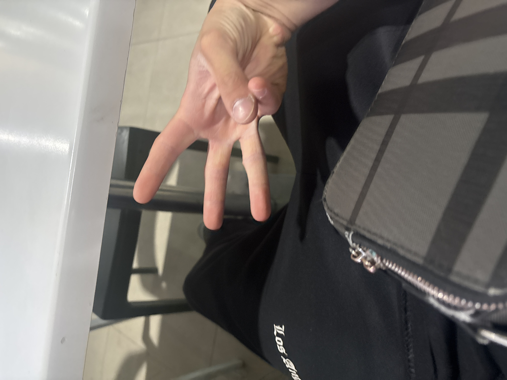 The image presents a close-up, slightly tilted view of a person's right hand, which is held up, extending the index, middle, and ring fingers while the thumb and pinky are folded. This gesture typically represents the number three. The hand has a light skin tone. To the right, a segment of a bag or purse is visible, featuring a distinct grey and black plaid or striped pattern and a silver zipper with a circular pull. The person is dressed in dark, likely black, casual clothing, possibly sweatpants, with stylized white text, appearing to read "Los Angeles," partially visible on the left thigh area. On the far left, a prominent white surface, possibly a table or wall, occupies a significant portion of the frame. In the background, dark, angular shapes suggest elements of furniture, such as chair or table legs, set against a lighter, undefined floor or wall. The scene appears to be indoors, illuminated by artificial lighting. The location context for this image is Yerevan, Armenia.