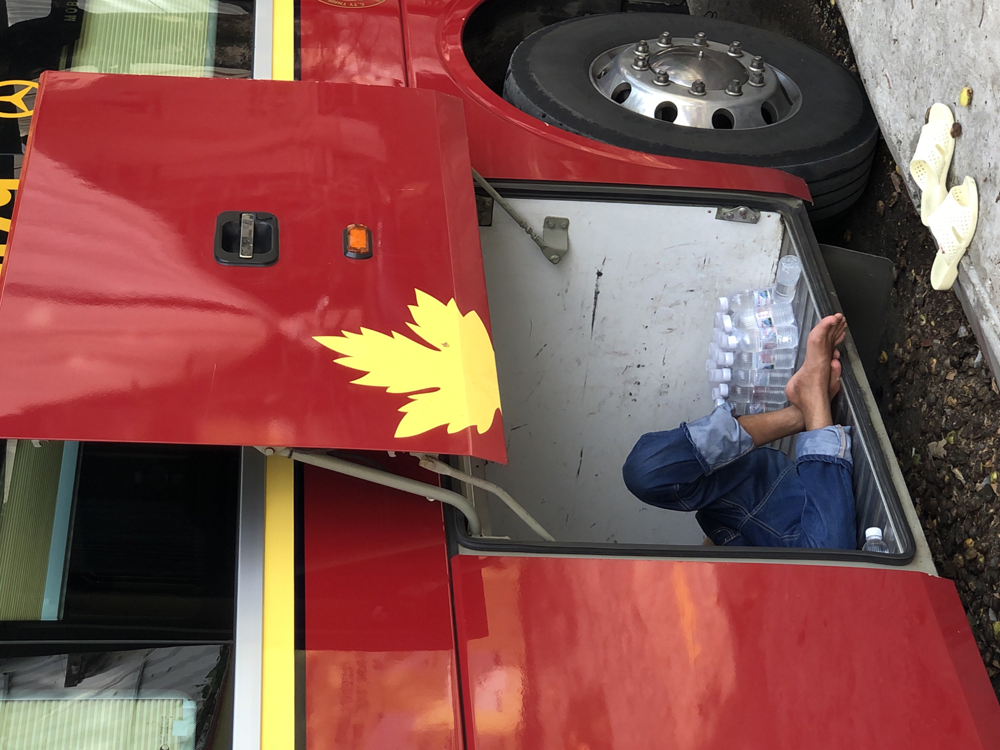A person in blue jeans is sitting in the open storage compartment of a red bus. The person's legs are visible, and they are bare from the knees down, with their feet resting on the floor of the compartment. There are several plastic water bottles lined up next to the person. Outside the bus, on the ground, a pair of yellow flip-flops and some debris are visible. The bus has a yellow stripe along its side and a yellow leaf design on the red exterior. The image was taken in Đà Nẵng, Vietnam.