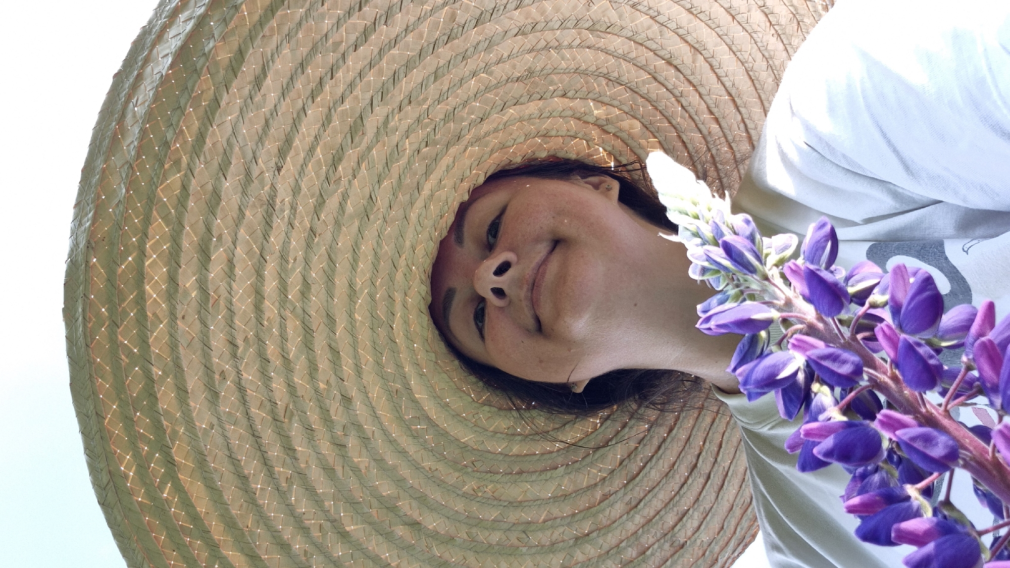 A low-angle, wide-shot photograph shows a person from the shoulders up, wearing a large, woven straw hat and a light-colored t-shirt. The person is holding a cluster of purple and white lupine flowers in the foreground, obscuring their chest. The hat takes up the majority of the frame, with the brim casting a shadow over the person's face. The person is looking upwards at the camera, with a slight smile. The background is a bright, overcast sky. The image does not provide any specific details about the location within the city of Новостройка, Russia.