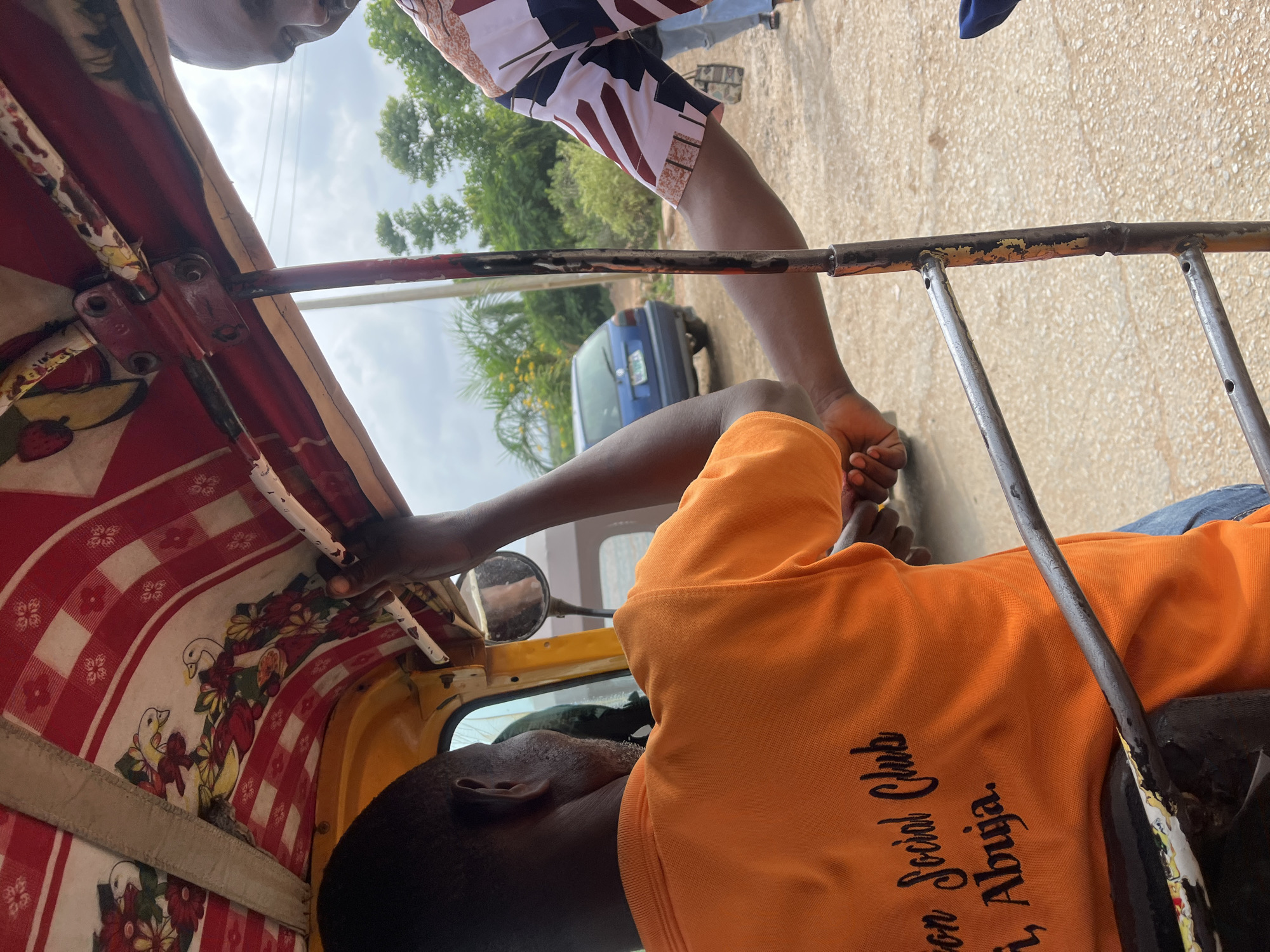 The image shows the interior of a yellow and red motorized rickshaw, a common form of transport in Kuchingoro, Nigeria. The driver, wearing an orange t-shirt with the text "Social Club, i, Abuja" visible, is seated and operating the vehicle. Another person, wearing a patterned shirt, is visible in the foreground, leaning towards the driver. The rickshaw's ceiling is decorated with a red and white checkered pattern and floral motifs. Outside the rickshaw, a blue car is parked on a dirt road, with lush green vegetation and trees in the background under a cloudy sky. The scene depicts everyday life and transportation in a Nigerian city.
