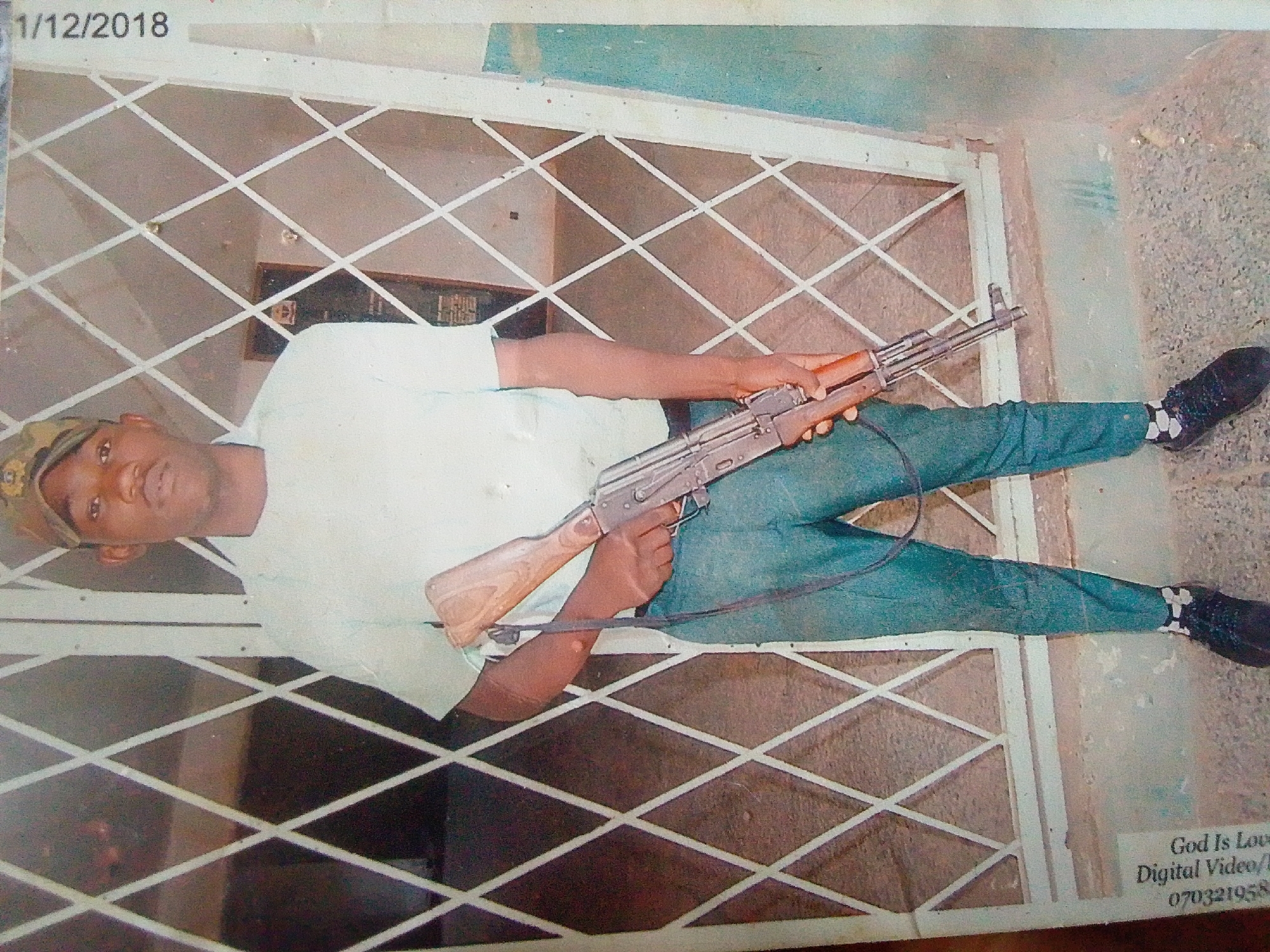 A young man in a light-colored shirt, dark pants, and a cap lies on his back in front of a white, diamond-patterned metal grate. He holds an assault rifle with a wooden stock and dark metal body. His left leg is bent, and his right leg is extended. The background appears to be a building wall with a framed picture visible through the grate. The image is dated "1/12/2018" in the upper left corner and has text in the lower right corner that reads "God Is Love Digital Video/ 070321958".