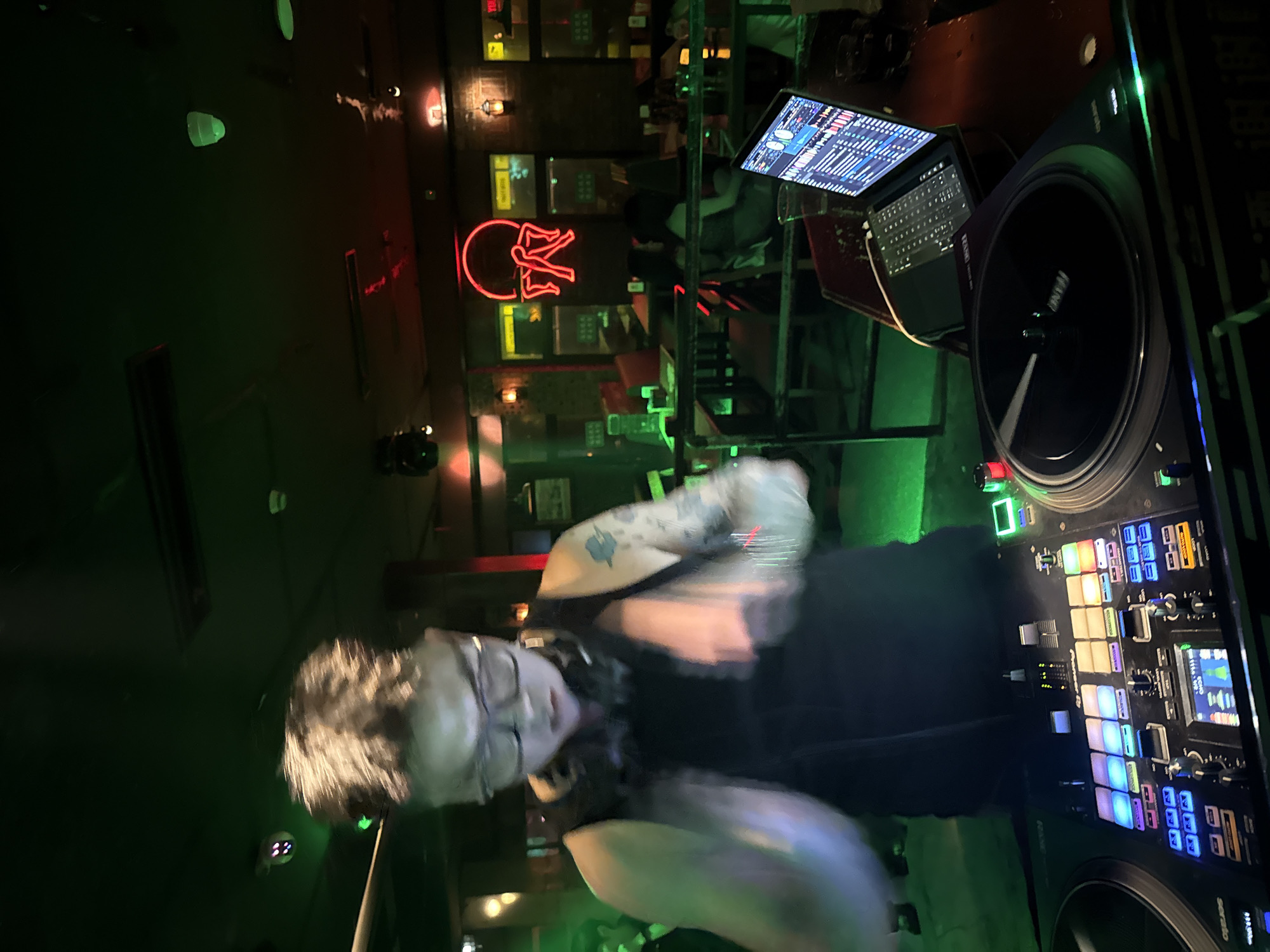 A DJ is seen actively performing in a dimly lit establishment, likely a nightclub or bar, in Liangjiang, China. The DJ, wearing glasses and a black tank top, has headphones around their neck and visible tattoos on their arms. They are blurred due to motion, suggesting they are engaged in mixing music. In front of the DJ is a DJ setup, including turntables, a mixer with numerous illuminated buttons and faders, and a laptop displaying music software. The environment is characterized by green and red lighting, with a neon sign featuring a red abstract figure visible in the background. Tables and chairs are set up in the background, with other patrons seated. The overall atmosphere appears to be one of nightlife and entertainment.