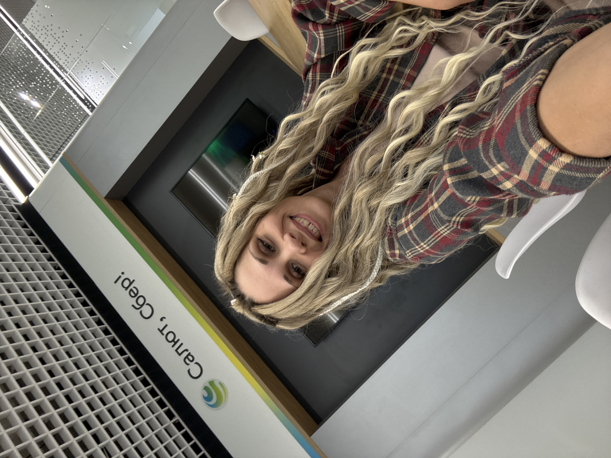A woman with long, blonde, curly hair is wearing a grey and red plaid flannel jacket in an indoor setting. She is smiling at the camera. Behind her is a wall with text in Russian, which includes "Сбер." The wall has a green and yellow stripe. To the left of the wall, there is a grid-like structure. The woman appears to be sitting, possibly in a chair, inside a Sberbank branch in Rusanovo, Russia.