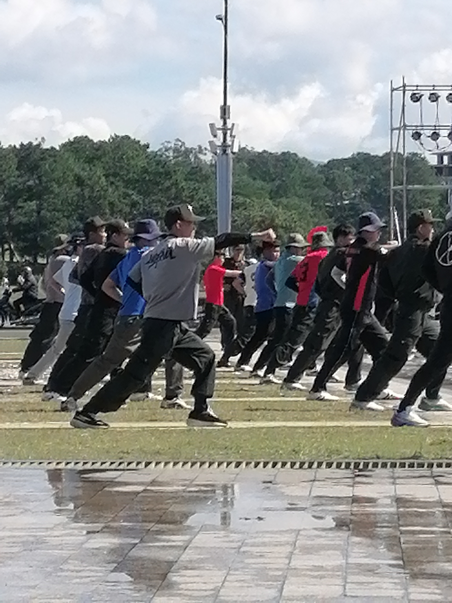 In Da Lat, Vietnam, a group of individuals, primarily young men, are captured in a dynamic, synchronized movement outdoors. They are arranged in a diagonal line, facing towards the left of the frame, and appear to be engaged in a form of group exercise or martial arts practice. Their bodies are angled, with arms extended forward and legs positioned in a lunging stance, suggesting a moment of intense physical activity.
The individuals are dressed in athletic attire, with a variety of colors and styles evident, including t-shirts, long-sleeved shirts, and trousers. Some are wearing baseball caps, adding to their casual and active appearance. The background consists of lush green trees, indicating a park or a natural setting within the city. Above the trees, a bright, overcast sky with scattered clouds provides a diffused light for the scene. Further in the background, metallic scaffolding structures and lighting equipment suggest the possibility of an outdoor event or performance taking place nearby. The foreground features a wet, tiled surface, possibly a pathway or plaza, reflecting the surrounding environment and adding a sense of depth to the image. The overall impression is one of focused energy and collective action within a pleasant, outdoor urban space.