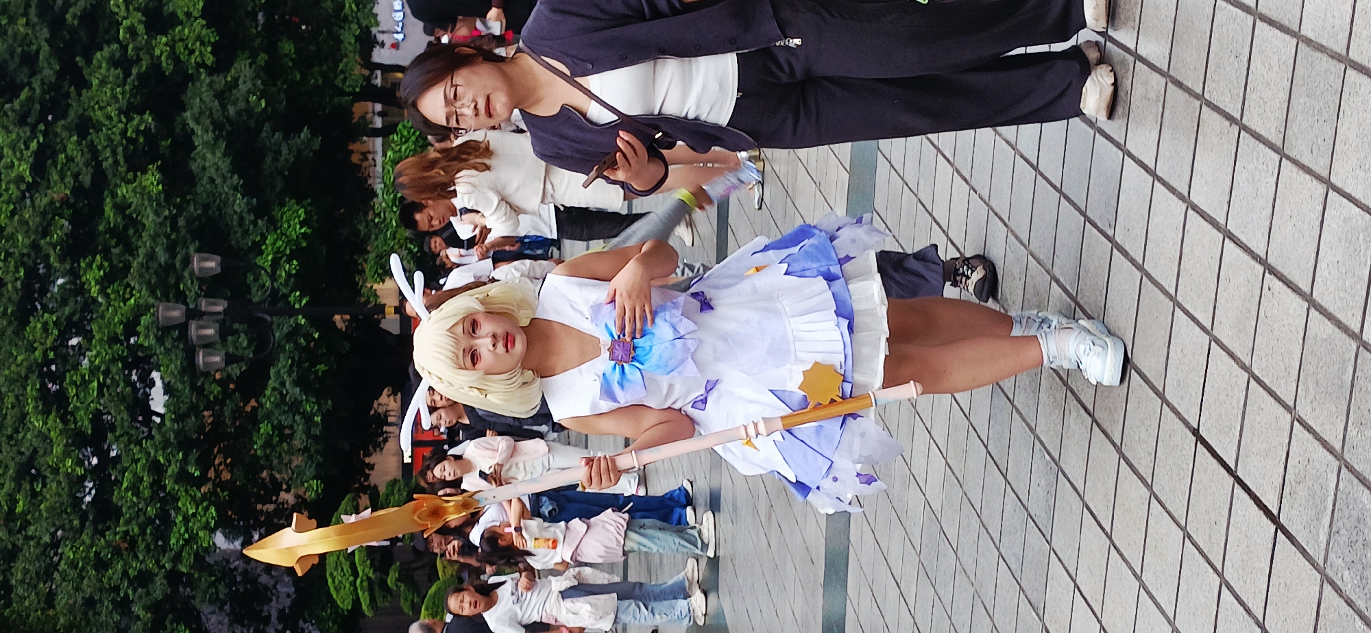 The photo shows a person dressed in a cosplay outfit in the city of Yuzhong District, China, evidenced by Chinese people present in the background. The cosplayer is wearing a white dress with purple and blue accents, a blond wig with white antlers, and red contact lenses. They are holding a golden prop that appears to be a staff or weapon. They are standing on a paved surface with a grid pattern, which indicates a public area like a plaza or park. In the background, several people stand behind the cosplayer, some of whom appear to be watching or interacting with the cosplayer, while others are engaged in their activities. Some trees are visible on the left side of the image, suggesting the scene is outdoors in a park or public garden.