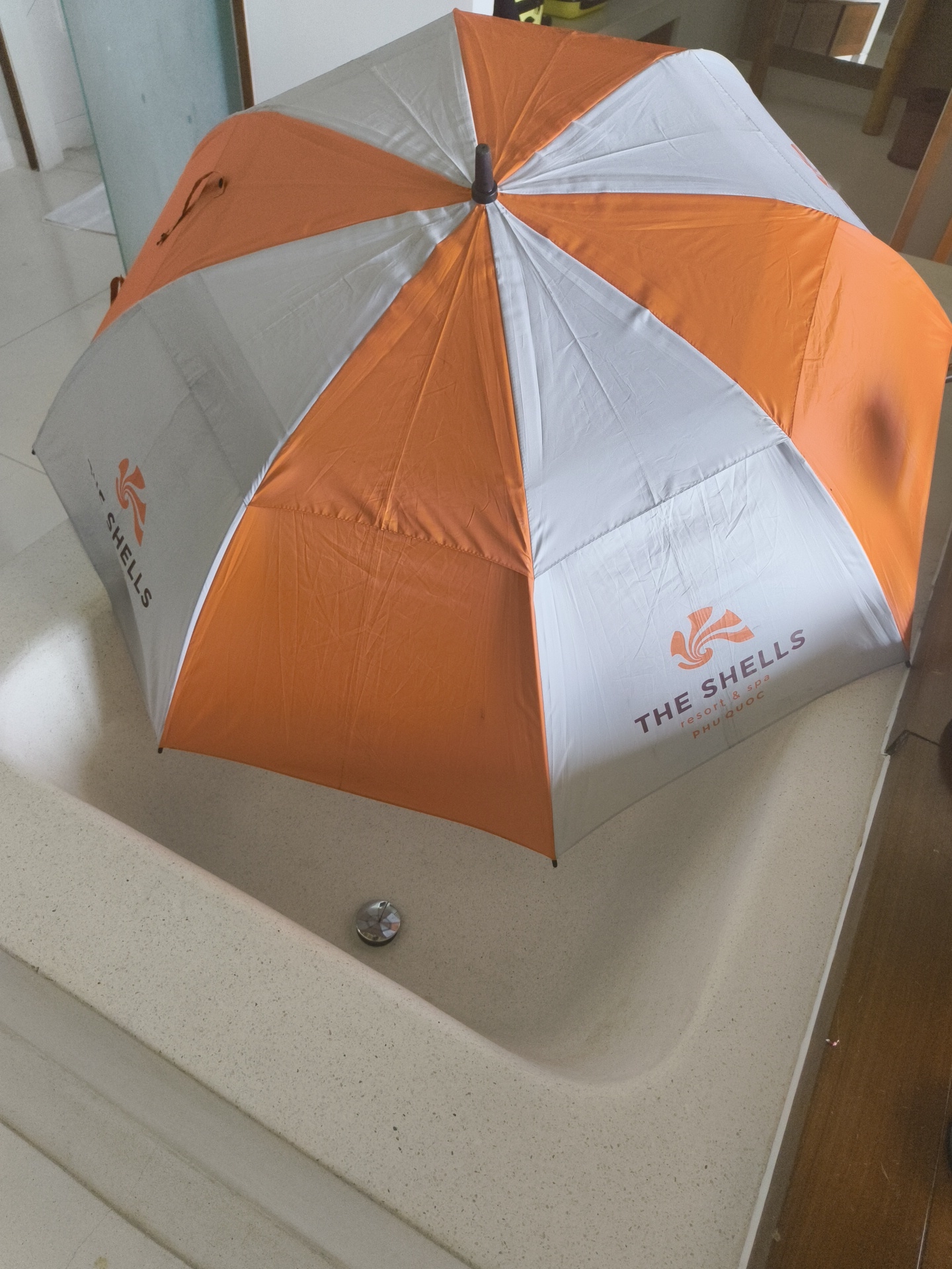 An orange and white umbrella is laid open inside a cream-colored bathtub. The umbrella has the logo "THE SHELLS resort & spa PHU QUOC" printed on one of its white panels. The bathtub appears to be made of a speckled material and has a drain visible in the center. The background shows a tiled floor, a glass partition, and some wooden furniture. The scene suggests a hotel room or resort setting in Phú Quốc, Vietnam.