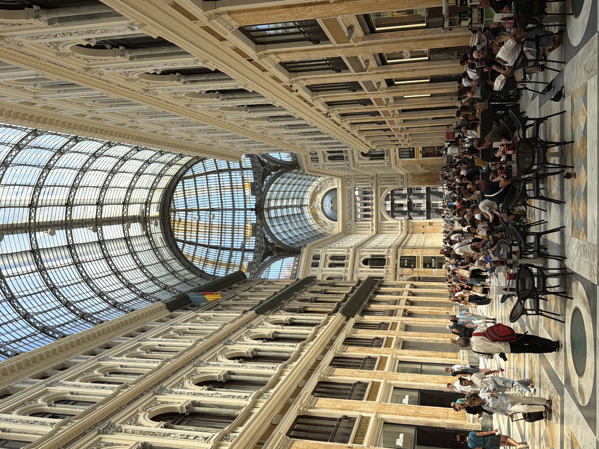 The image depicts the Galleria Umberto I, a public shopping gallery in Naples, Italy. It is a bustling scene with numerous people walking and seated at tables. The architecture of the gallery is a prominent feature, with ornate facades lining a long, wide walkway covered by a large glass and iron dome. Natural light streams in through the dome, illuminating the space.
In the foreground, several tables and chairs are set up for outdoor dining, with patrons enjoying drinks and conversation. A few pigeons are visible on the patterned marble floor. The middle ground is filled with a crowd of people of various ages, dressed in casual attire, moving in different directions. Some are looking at shops, while others are engaged in conversation.
The background reveals the further extent of the gallery, with its intricate architectural details and the continued presence of people. The overall atmosphere is lively and energetic, suggesting a popular public space within the city. The time of day appears to be daytime, indicated by the bright lighting. The floor is a decorative marble pattern with circular inlays. Storefronts with signage are visible along the sides of the gallery, contributing to the commercial aspect of the location.